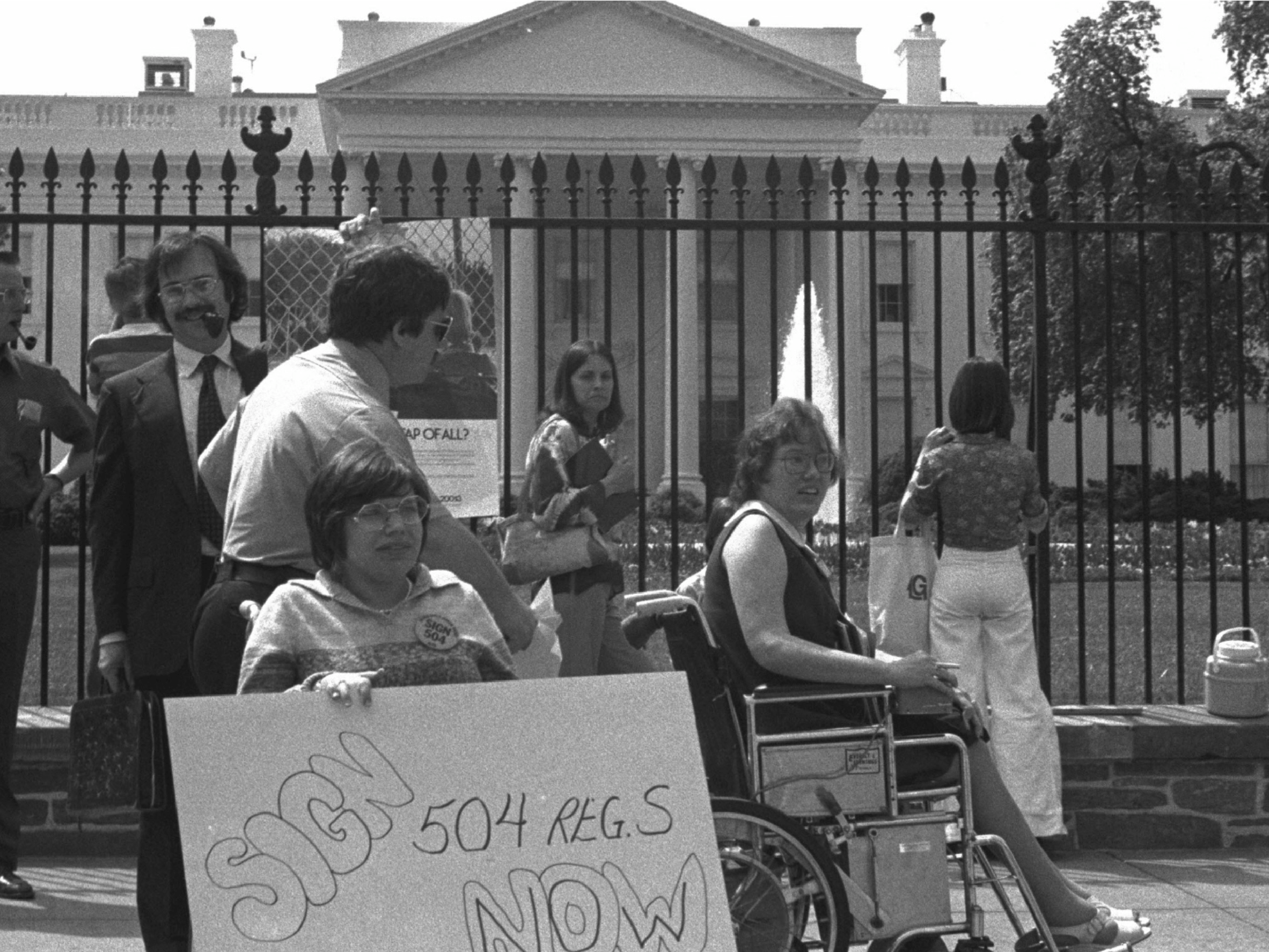 Heumann is pictured in her wheelchair in front of the White House, holding a sign that reads “Sign 504 Now.”