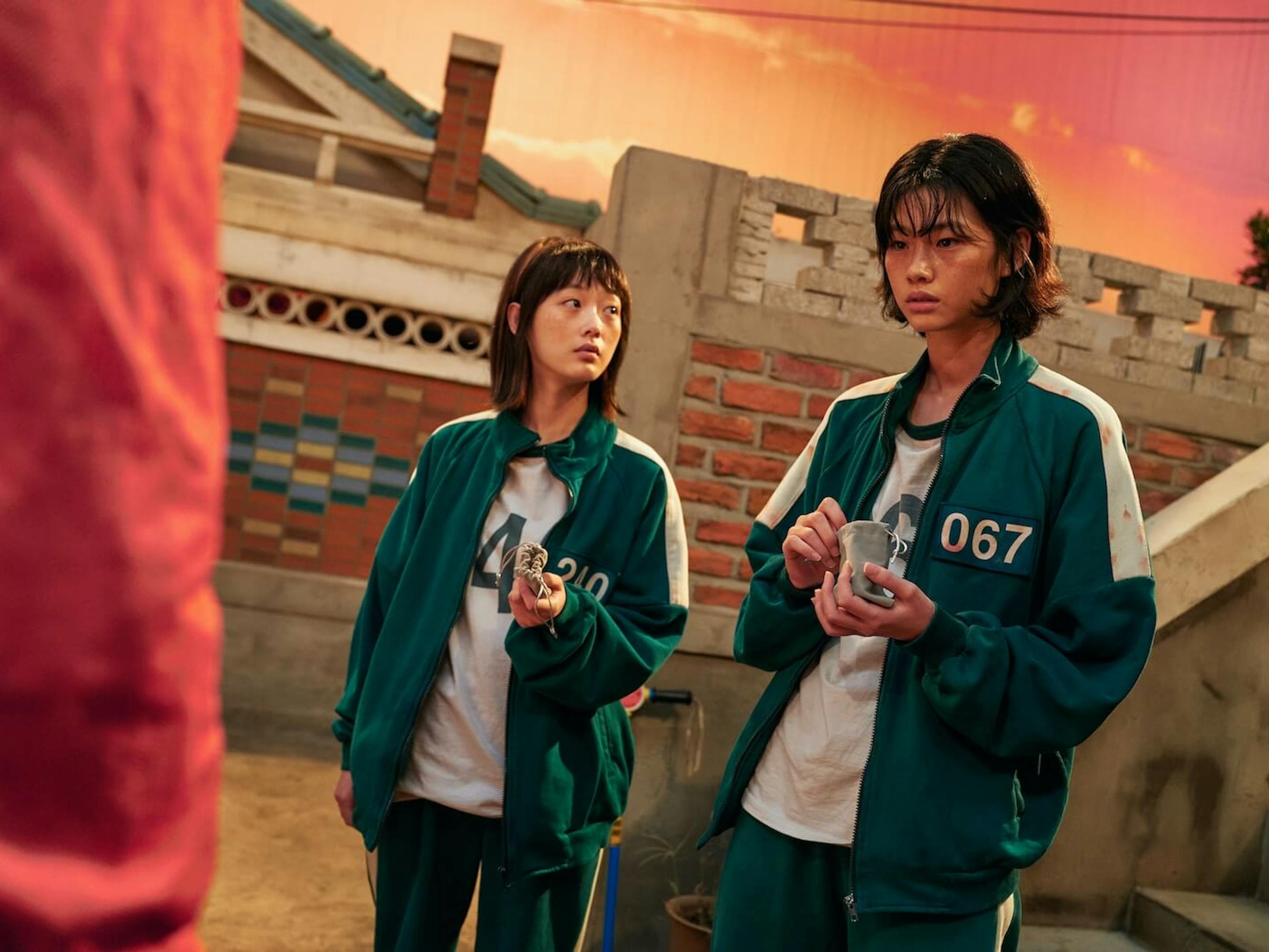 Ji-yeong (Lee You-mi) and Kang Sae-byeok (Jung Ho-yeon) stand together picking something out of a small leather bag. Their green tracksuits are unzipped and they look sweaty and afraid. In the background is a brick wall and a red and orange sky.