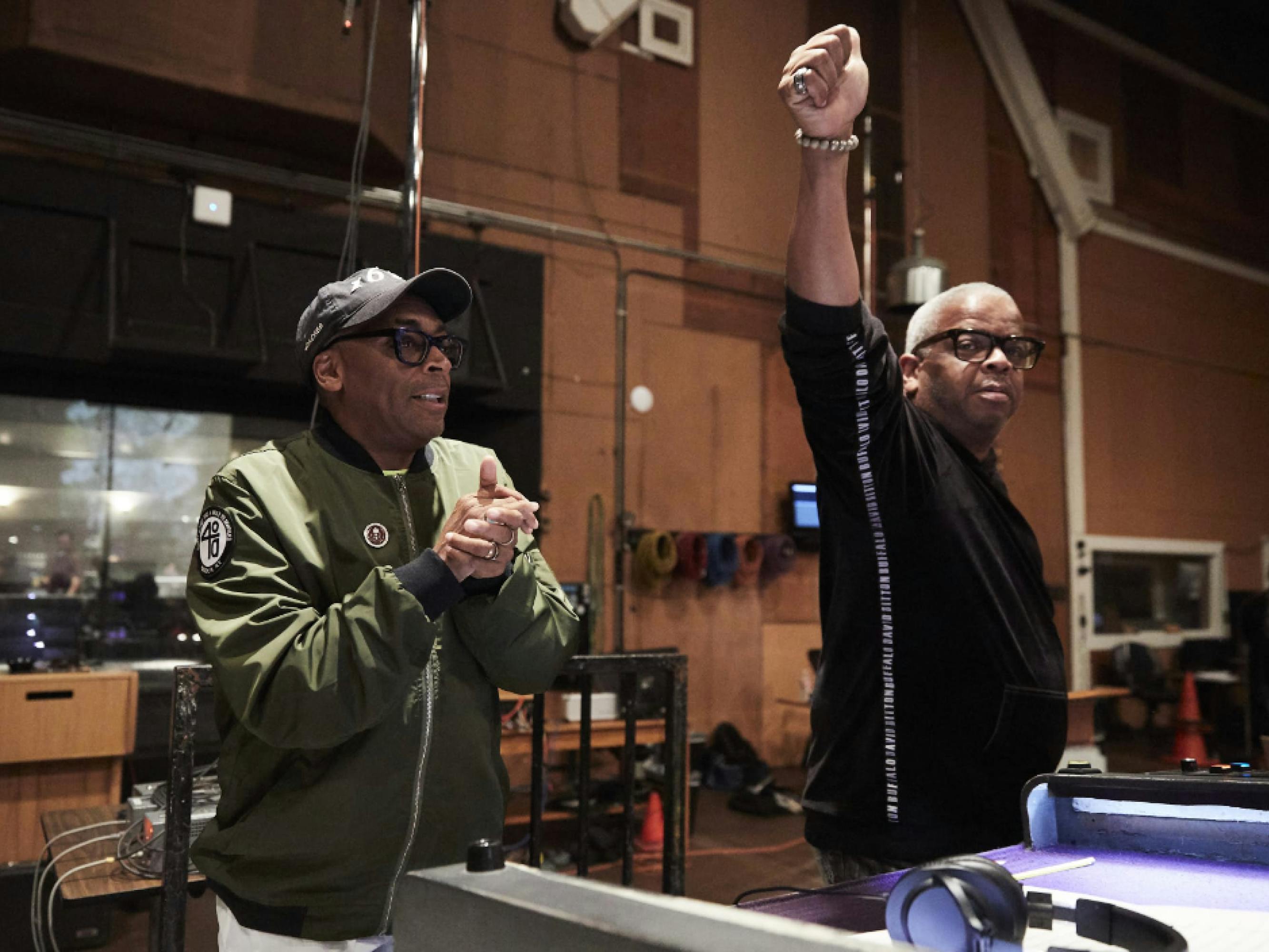 A photograph of Spike Lee and Terence Blanchard in the studio. The room is full of recording equipment. In the foreground is a pair of headphones. Lee has his hands clasped together and wears a black cap and green bomber jacket. Blanchard stands next to him, one fist raised in the air.