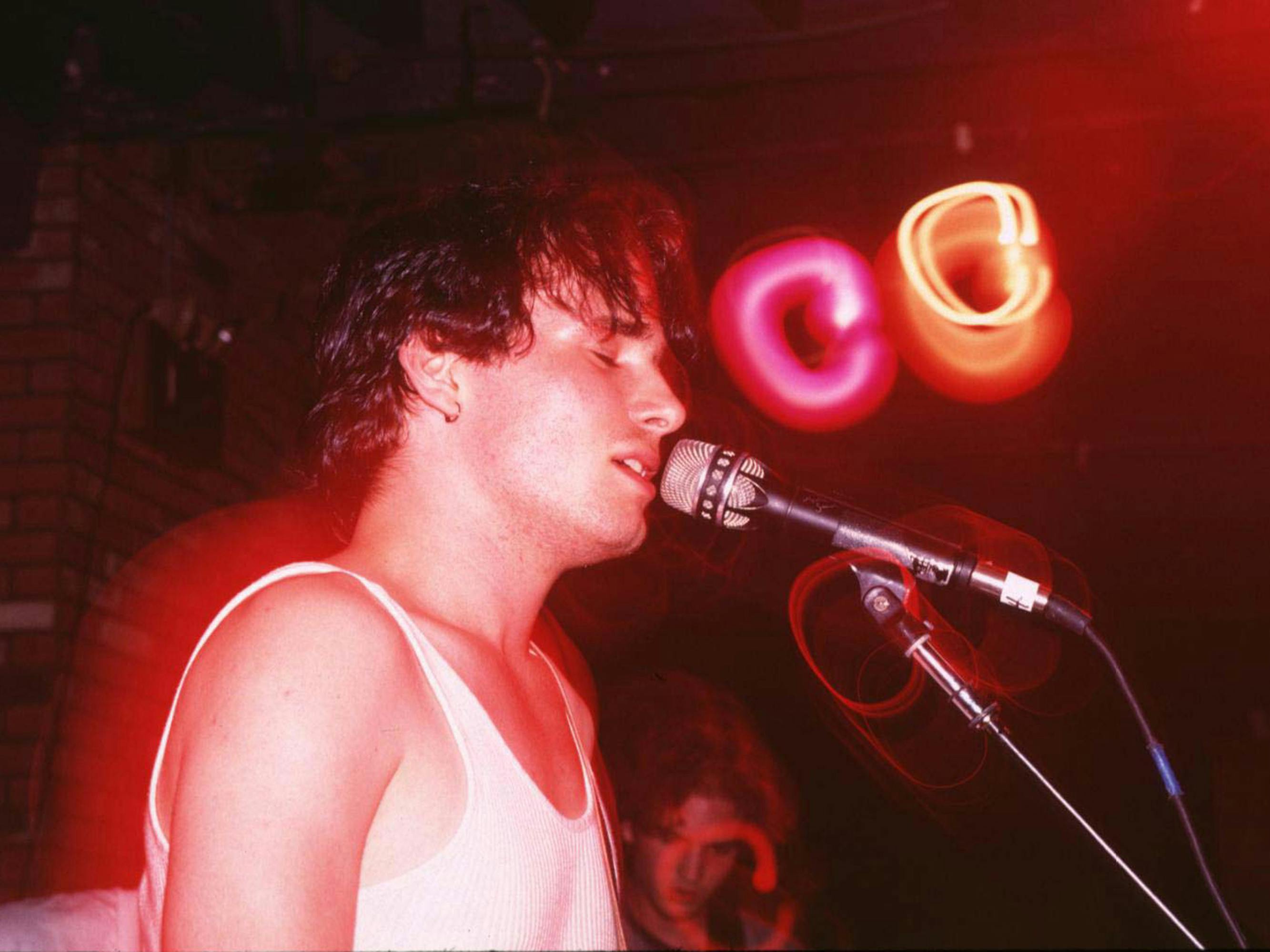 Jeff Buckley sings into a microphone earnestly, lit by red and yellow lights.