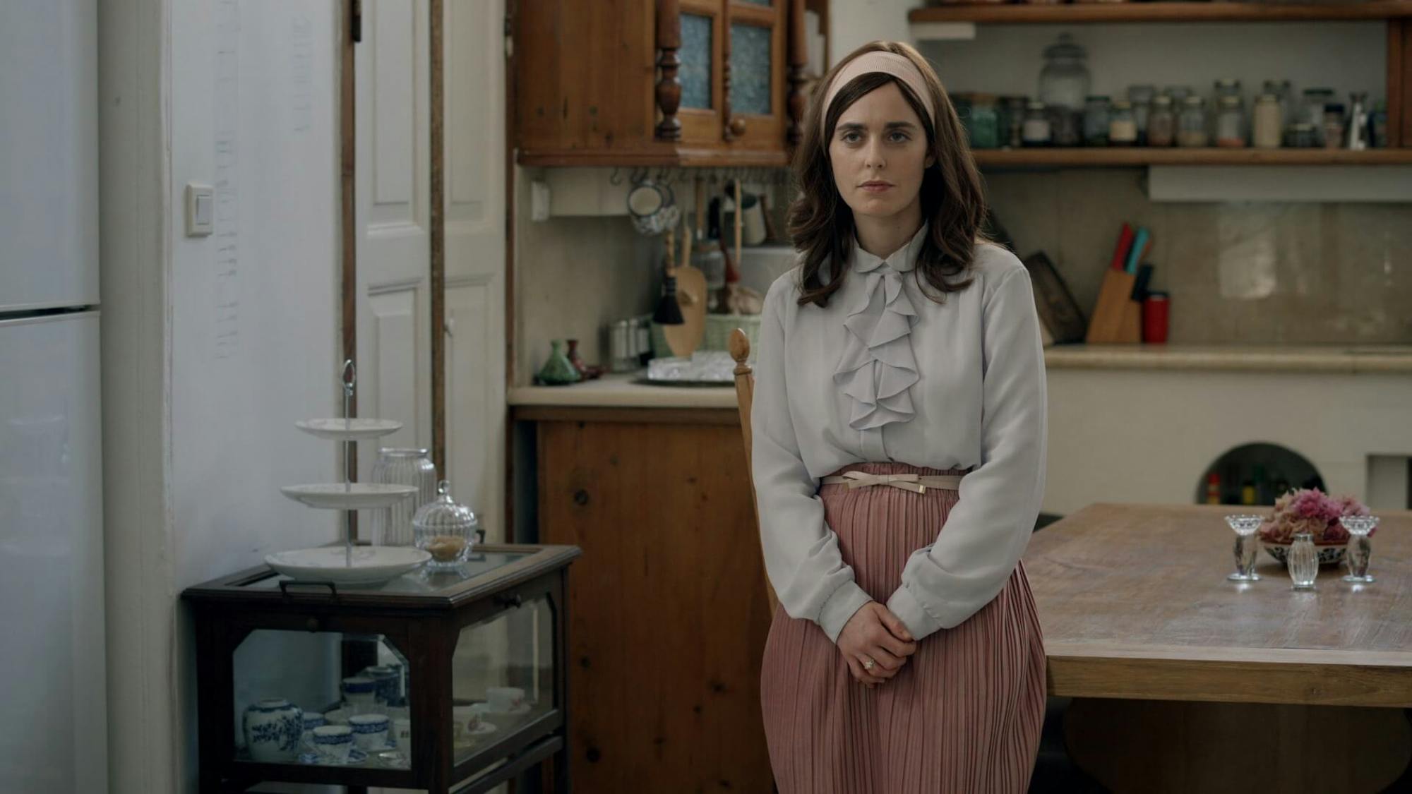 Libbi Shtisel (Hadas Yaron) stands in a kitchen looking prim and proper. She wears a pink skirt, pink headband, blue blouse, and small white belt. She crosses her arms in front of her body. In the background is a pristine kitchen.