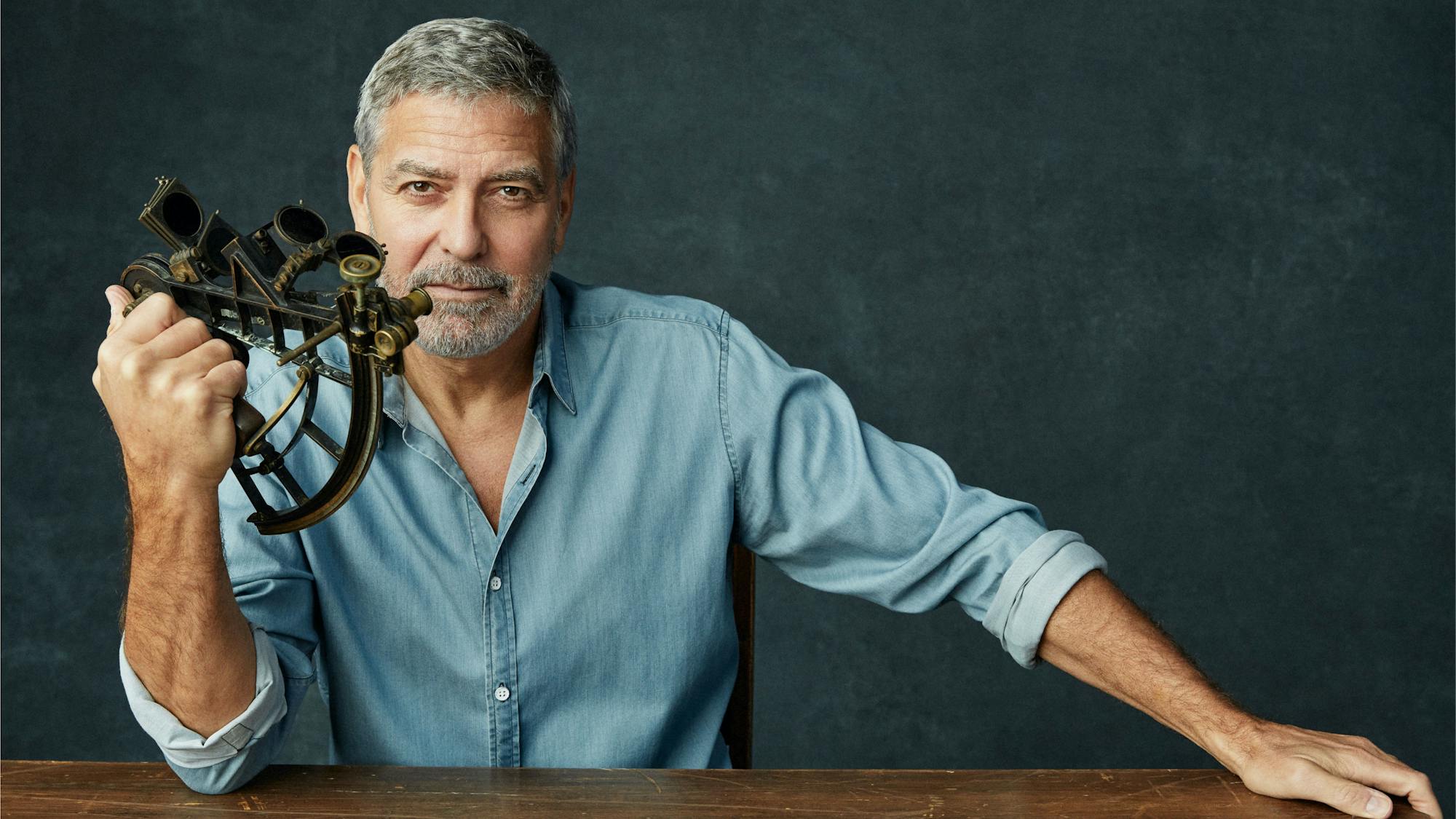 George Clooney sits in front of a dark wood table, wearing a light-blue denim shirt, sleeves rolled up, holding a sextant.