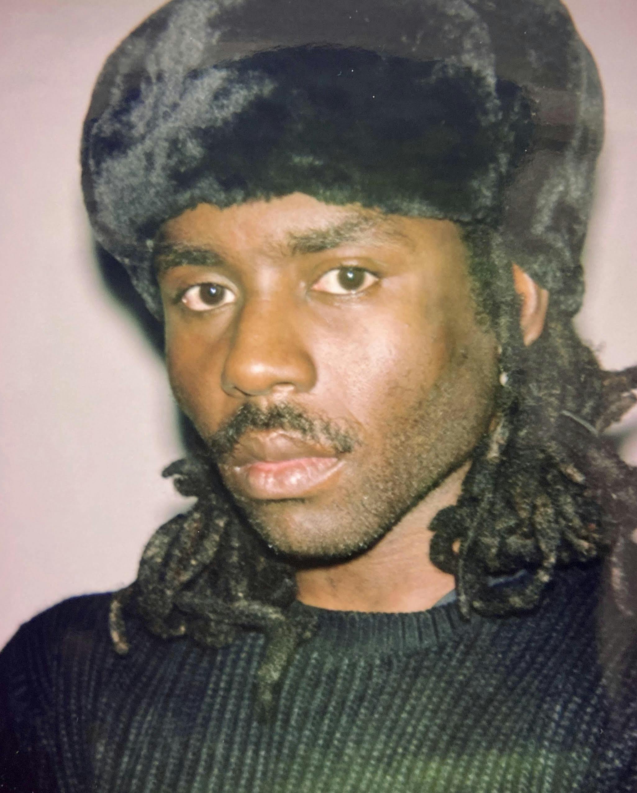 Dev Hynes wears a dark shirt and furry dark hat. He has a mustache and hair to his shoulders.