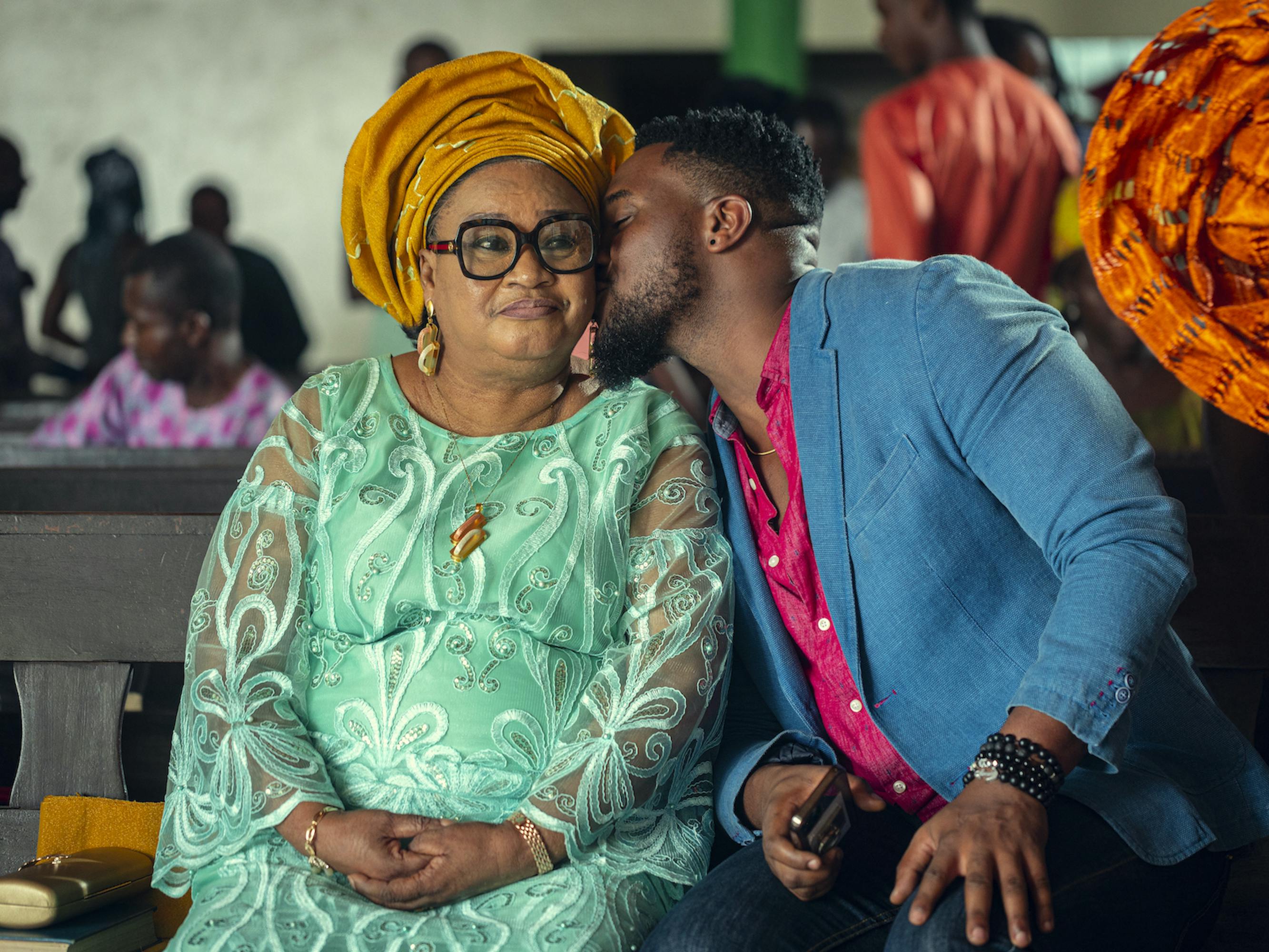 Rachel Oniga and Kunle Remi sit together. Oniga wears a beautiful turquoise dress and a yellow headwrap. Remi wears a blue blazer, hot pink shirt, and dark pants as he leans in to kiss Oniga on the cheek. 