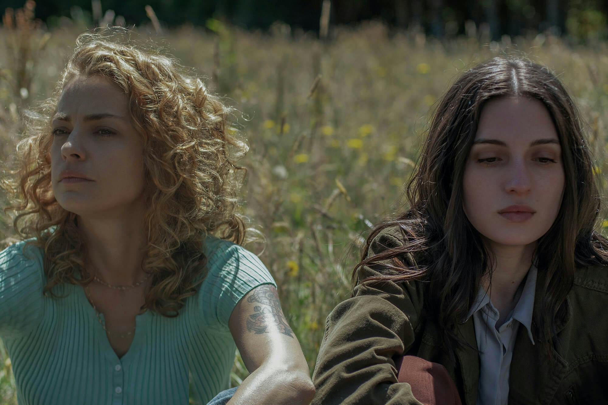Carola (Dolores Fonzi) y Amanda (María Valverde) se sientan en un campo de flores silvestres y pasto. Carola usa una camisa abotonada color turquesa y Amanda una chaqueta de color oscuro.
