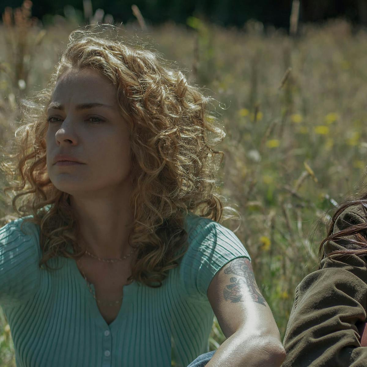 Carola (Dolores Fonzi) y Amanda (María Valverde) se sientan en un campo de flores silvestres y pasto. Carola usa una camisa abotonada color turquesa y Amanda una chaqueta de color oscuro.