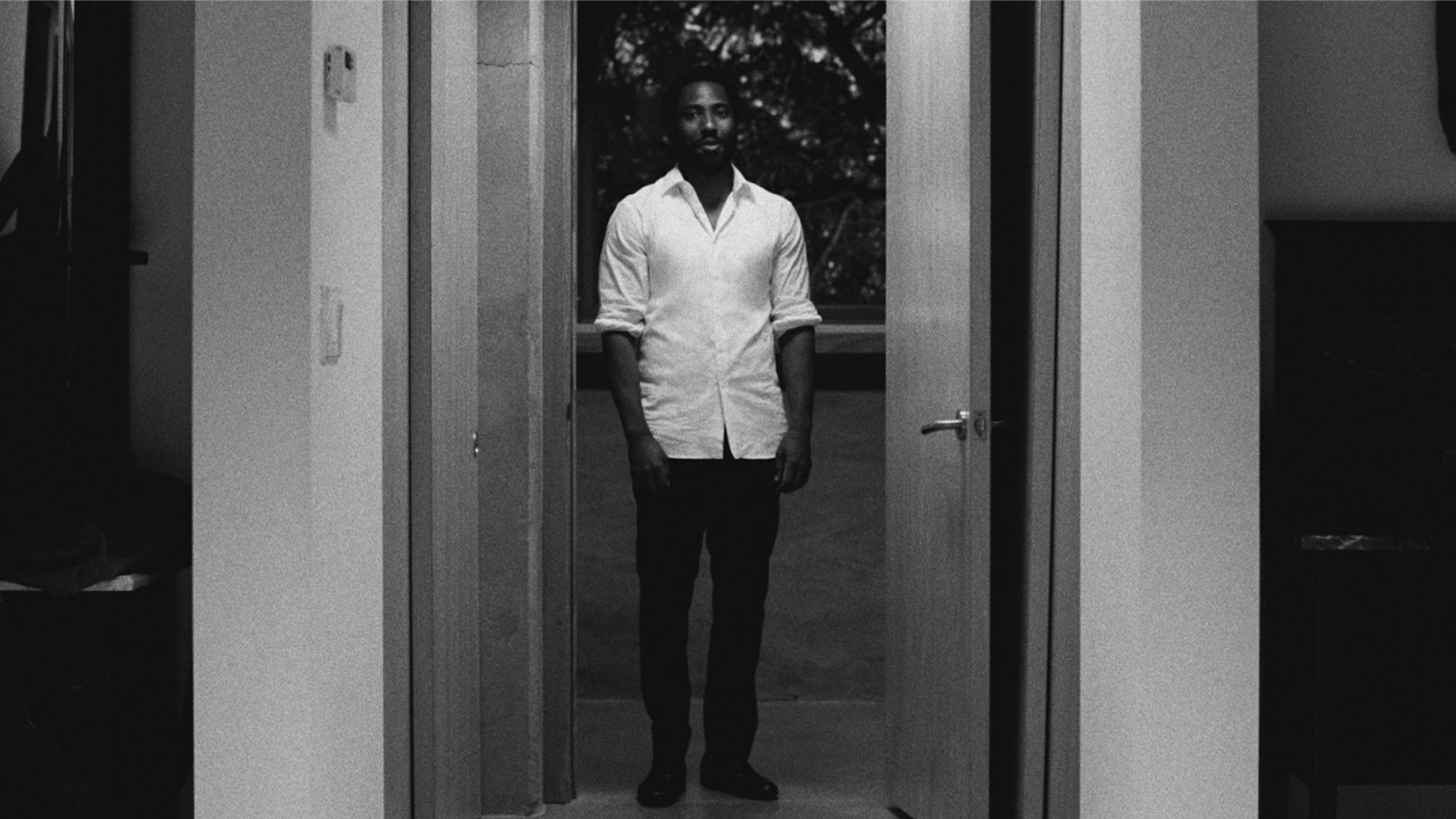Malcolm framed in a doorway, wearing a white shirt with the sleeves rolled up.