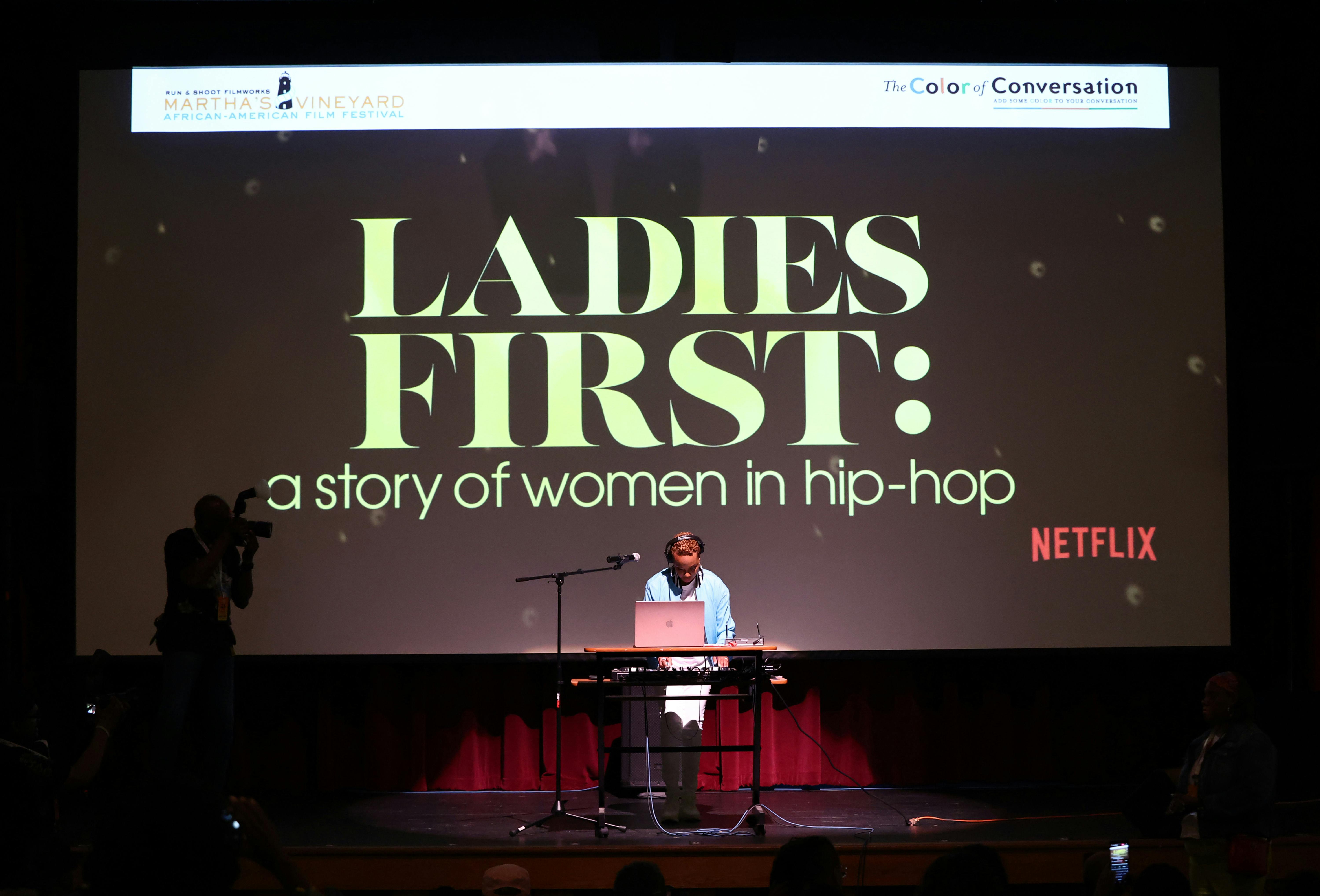 A woman appears to DJ from a laptop onstage in front of a projection of "LADIES FIRST: a story of women in hip-hop"
