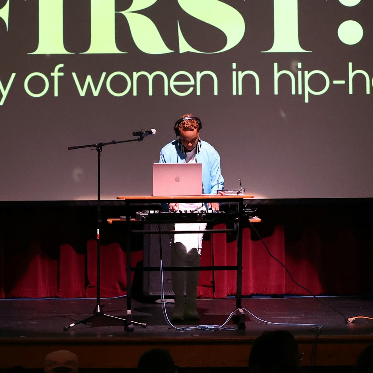 A woman appears to DJ from a laptop onstage in front of a projection of "LADIES FIRST: a story of women in hip-hop"