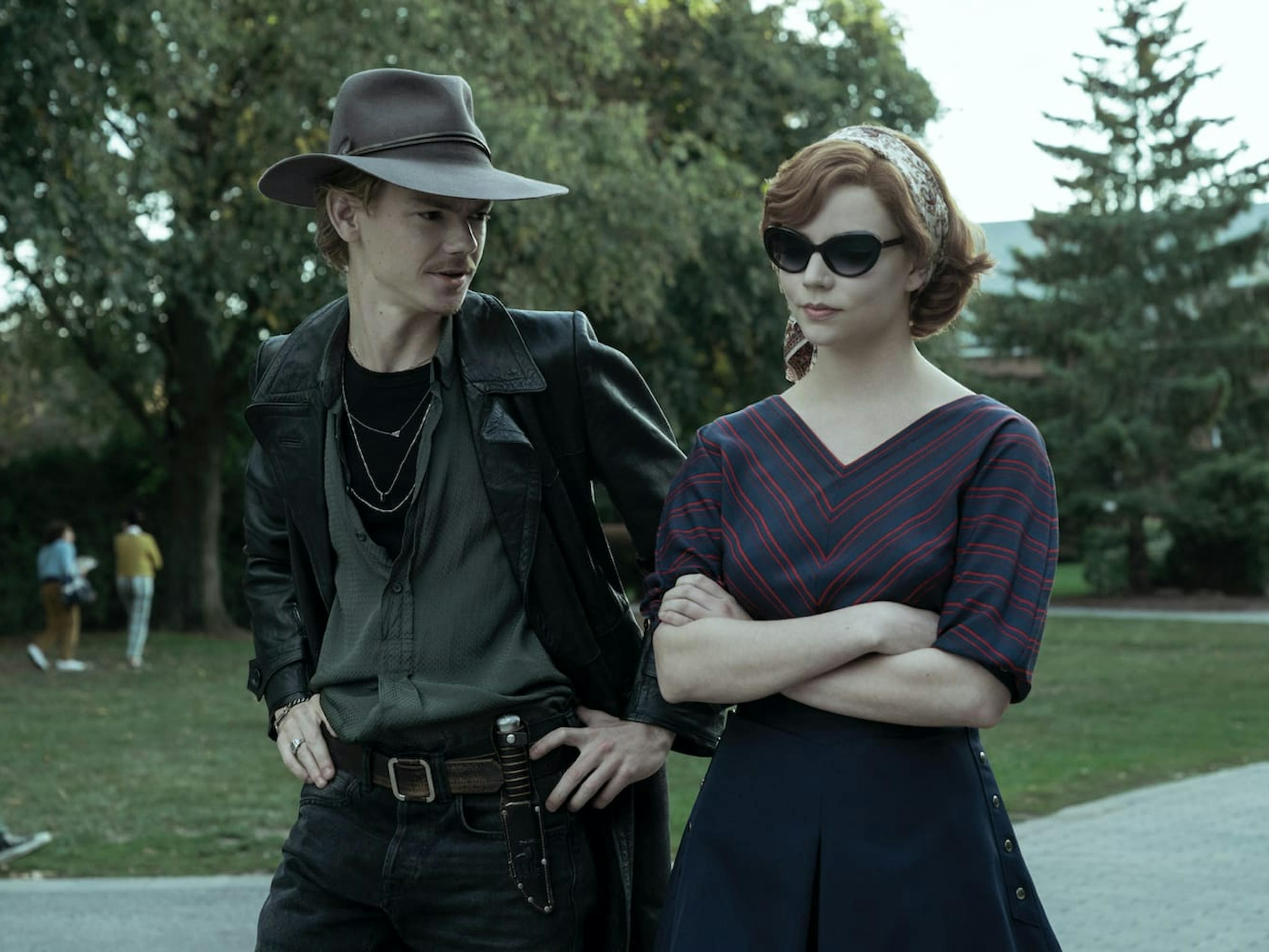 Benny Watts (Thomas Brodie-Sangster) and Beth Harmon (Anya Taylor-Joy stand together outside. Benny wears a leather jacket and wide brimmed hat. Beth wears a striped navy and red shirt and dark sunglasses.