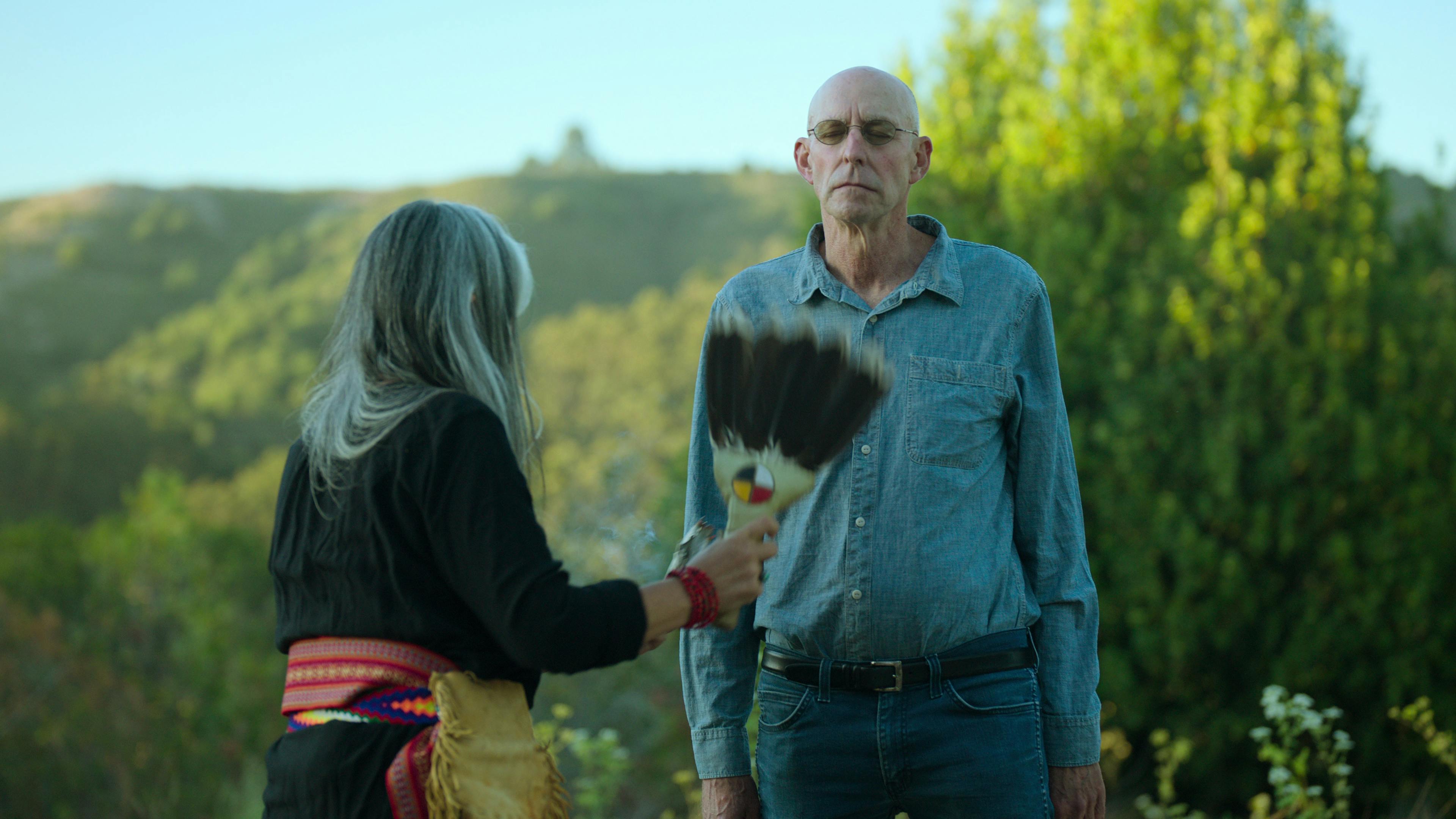 Michael Pollan participates in a tobacco ceremony. Behind him is a bright blue sky and green trees.