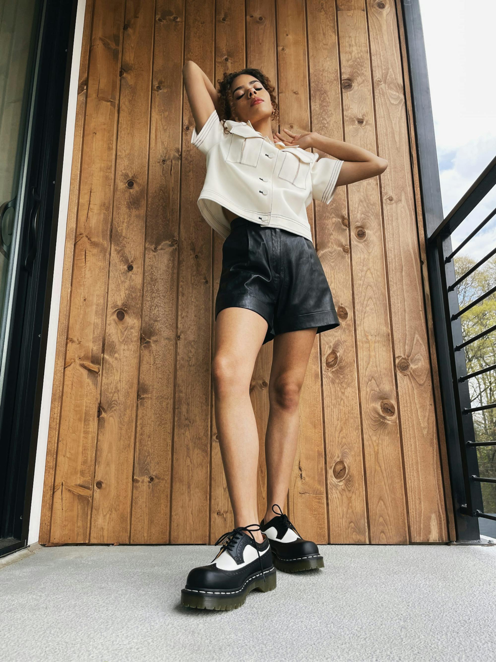 Gentry wears a white blouse with chunky stitching, leather shorts, and black-and-white platform brogue shoes. Her arms are perched above her head, and she stands in front of a wood panelled wall.