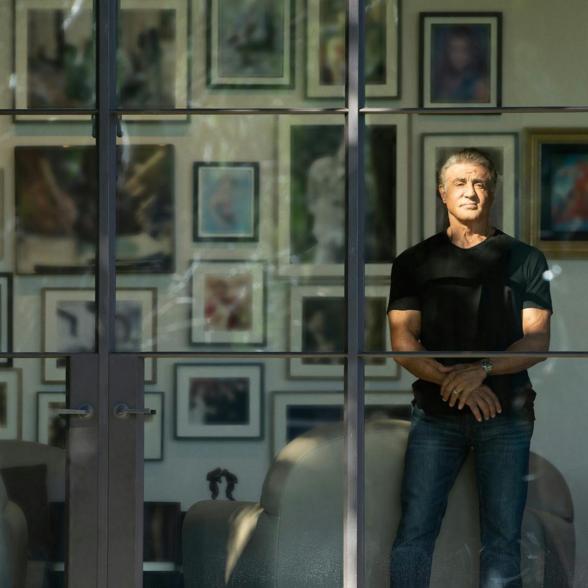Sylvester Stallone wears jeans and a black shirt and stands in front of a large window. 