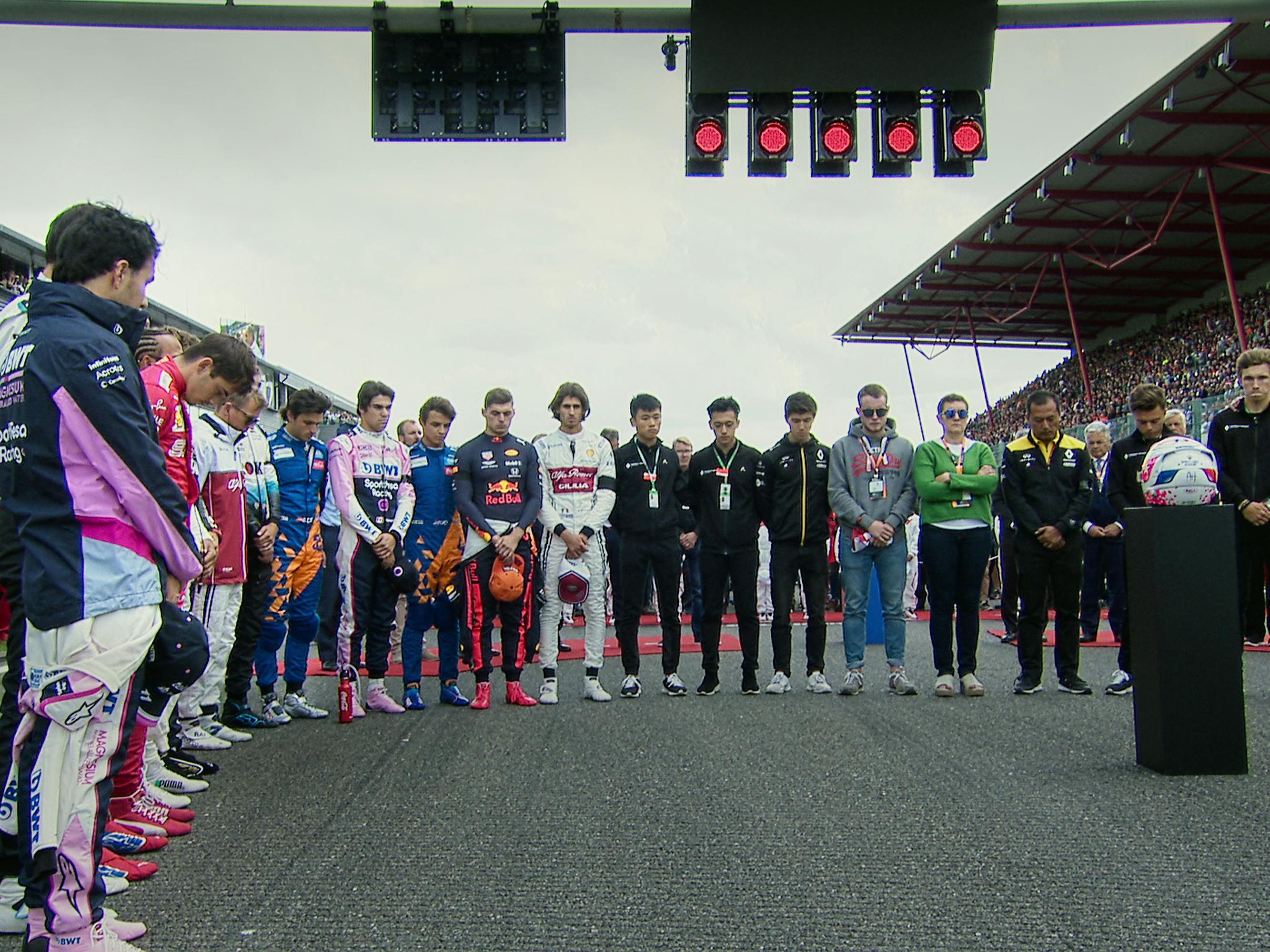 Drivers and the family of Anthoine Hubert stand around a pedestal and hold a minute of silence. Atop the pedestal is the helmet of Anthoine Hubert who had recently died in a crash during a race.