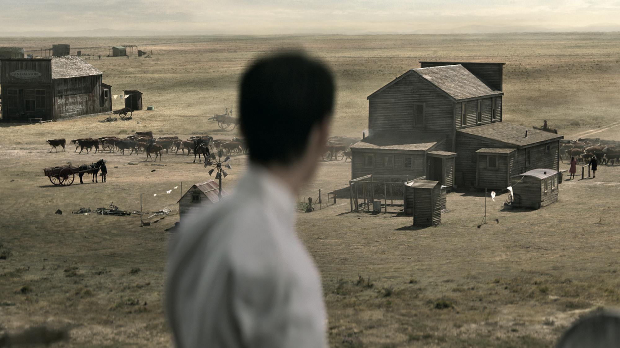 Kodi Smit-McPhee stands with his back to the camera in this blurry shot. There are horses and buildings dotting the dusty ground in front of him