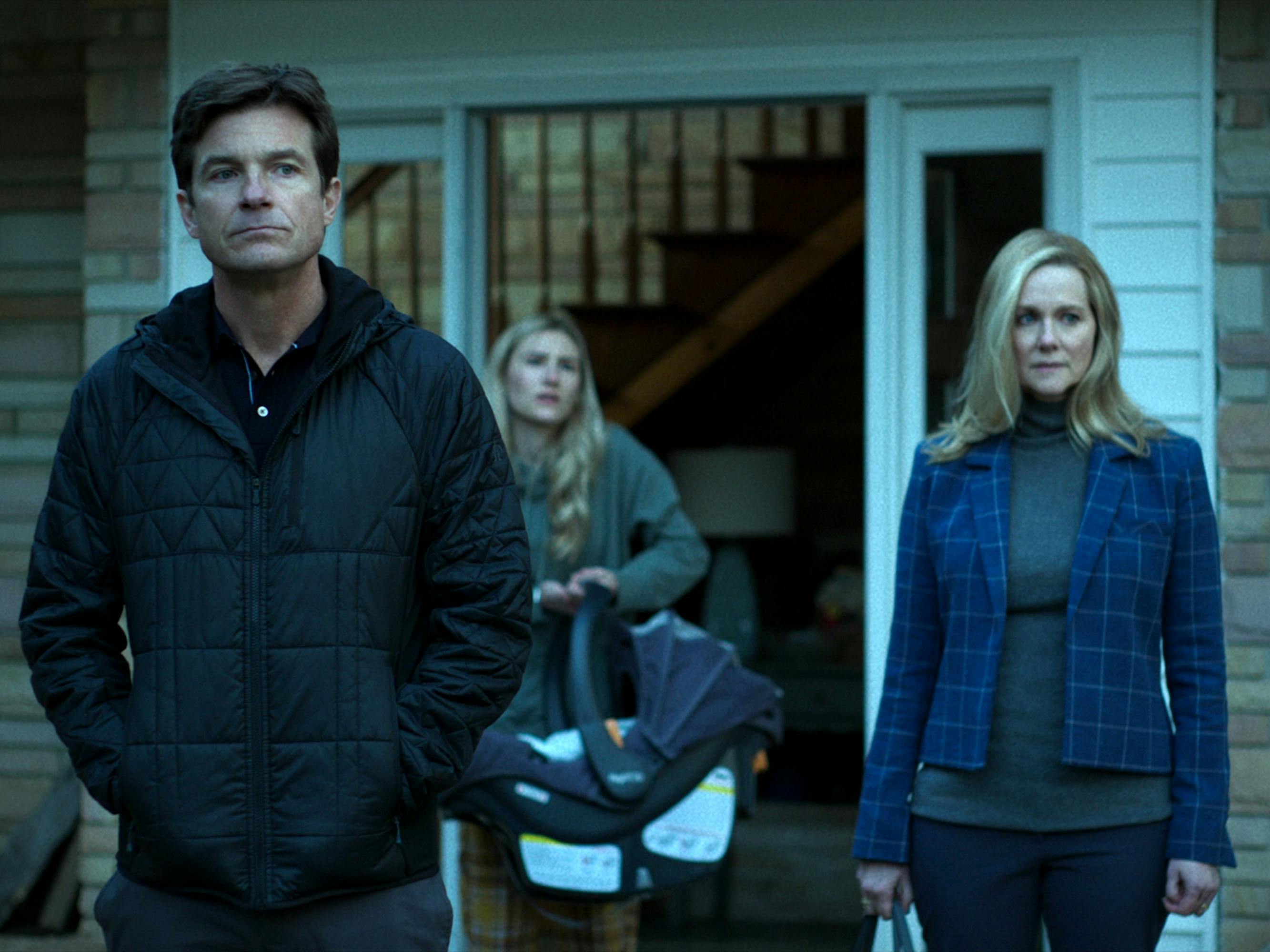 Jason Bateman and Laura Linney stand outside their house. Bateman wears a black puffer jacket and Linney wears a blue plaid jacket. 