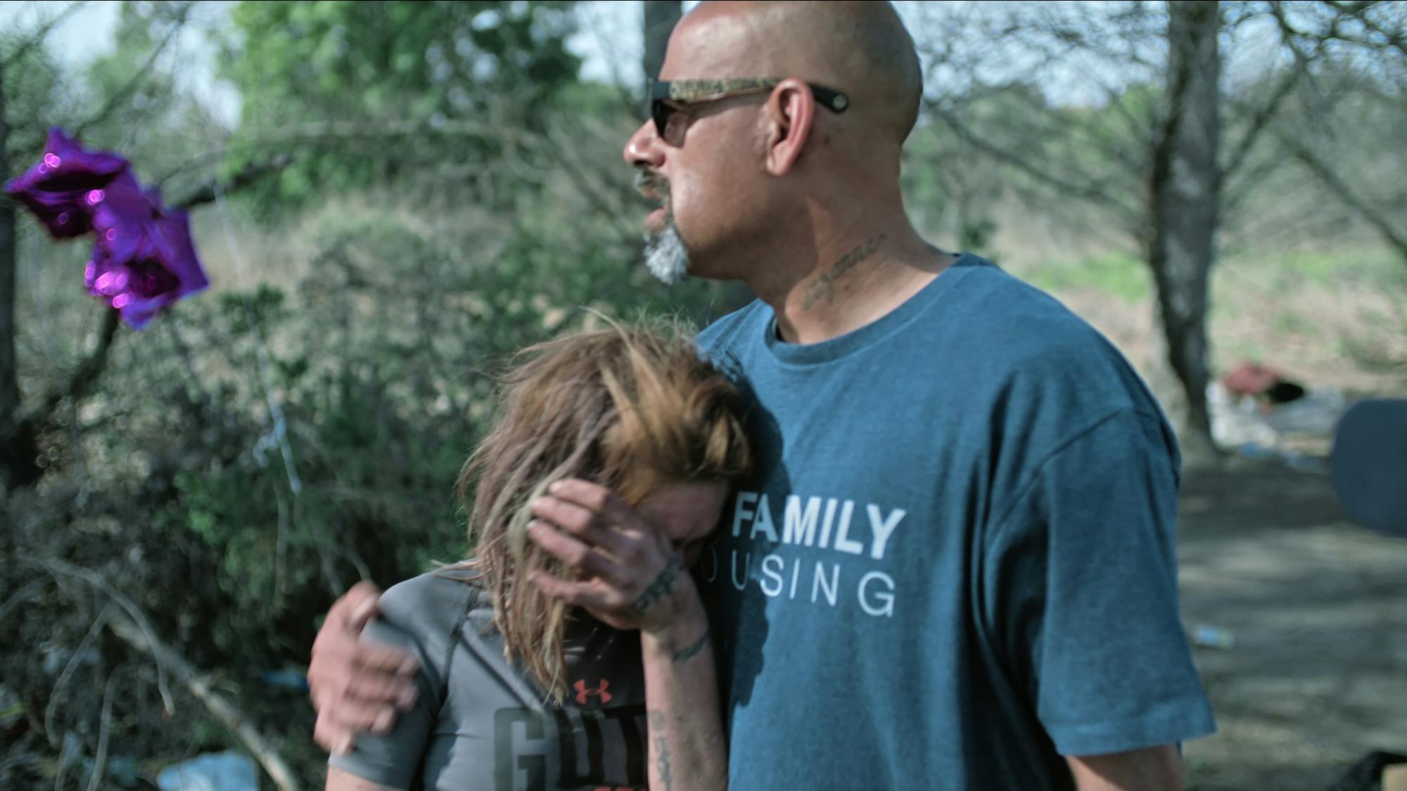 Patricia Wilcox rests her head against a man’s chest in this outdoor shot. She wears a grey t-shirt and wipes her eyes. He wears a grey shirt and sunglasses.