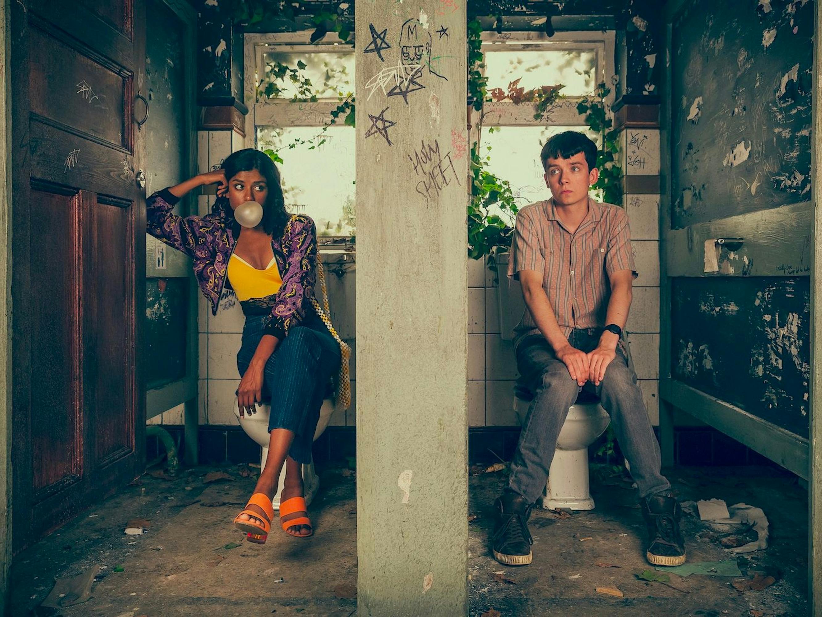 Olivia Hanan (Simone Ashley) and Otis Milburn (Asa Butterfield) in Sex Education. The two characters sit in the abandoned toilets. Ashly wears a chic outfit and blows a massive bubble. Butterfield wears dark jeans and a buttoned down shirt.