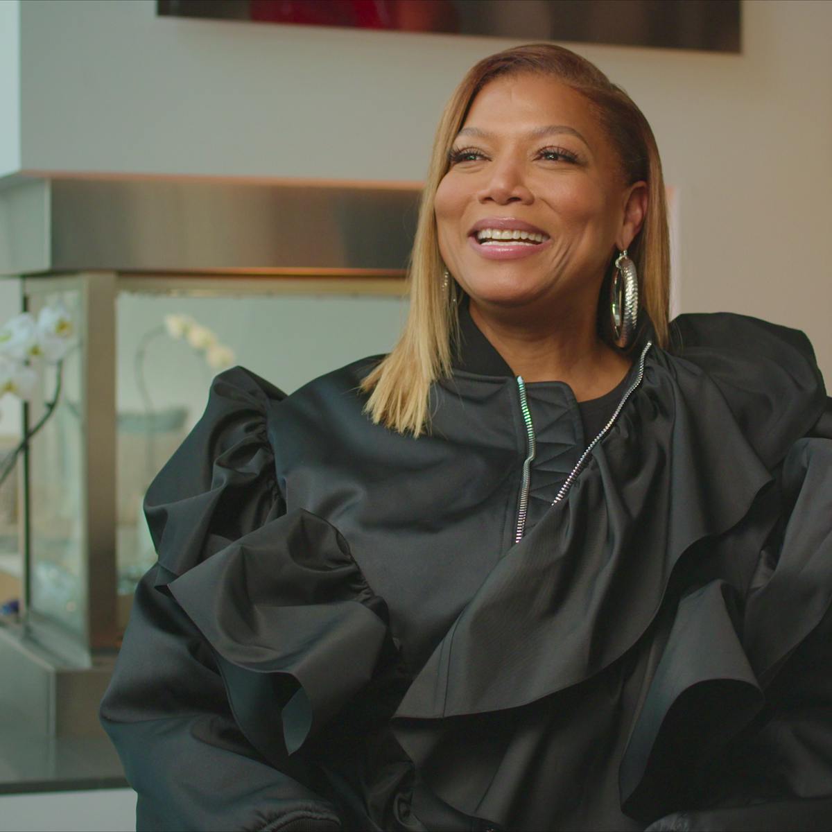 Queen Latifah wears a zippered, ruffled, black top and huge silver hoops. She smiles and looks at something off-camera. 
