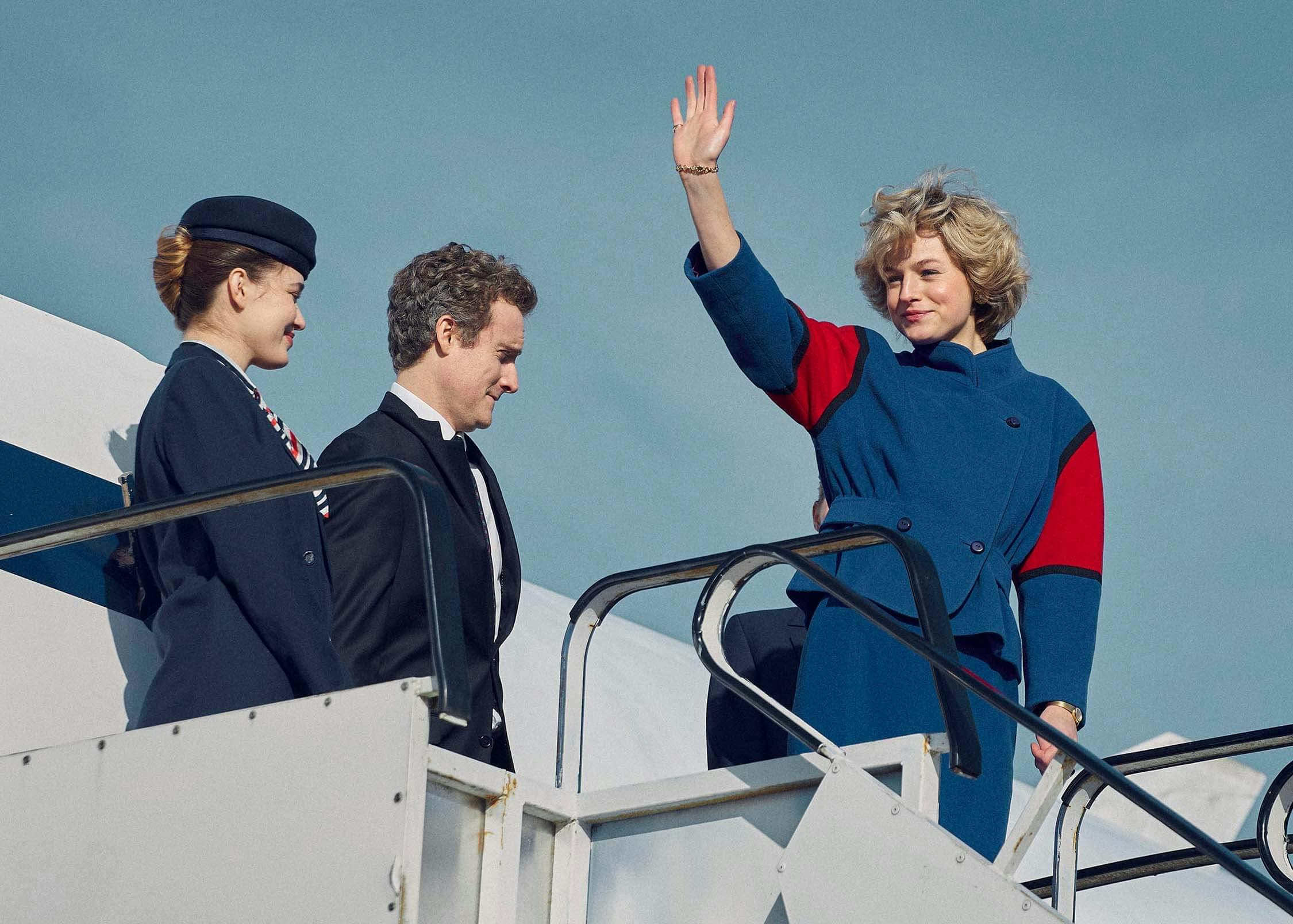 Diana (Emma Corrin) wears a red and blue suit as she exits a plane. Flanked by a man and a woman in dark suits, she waves to someone offscreen.