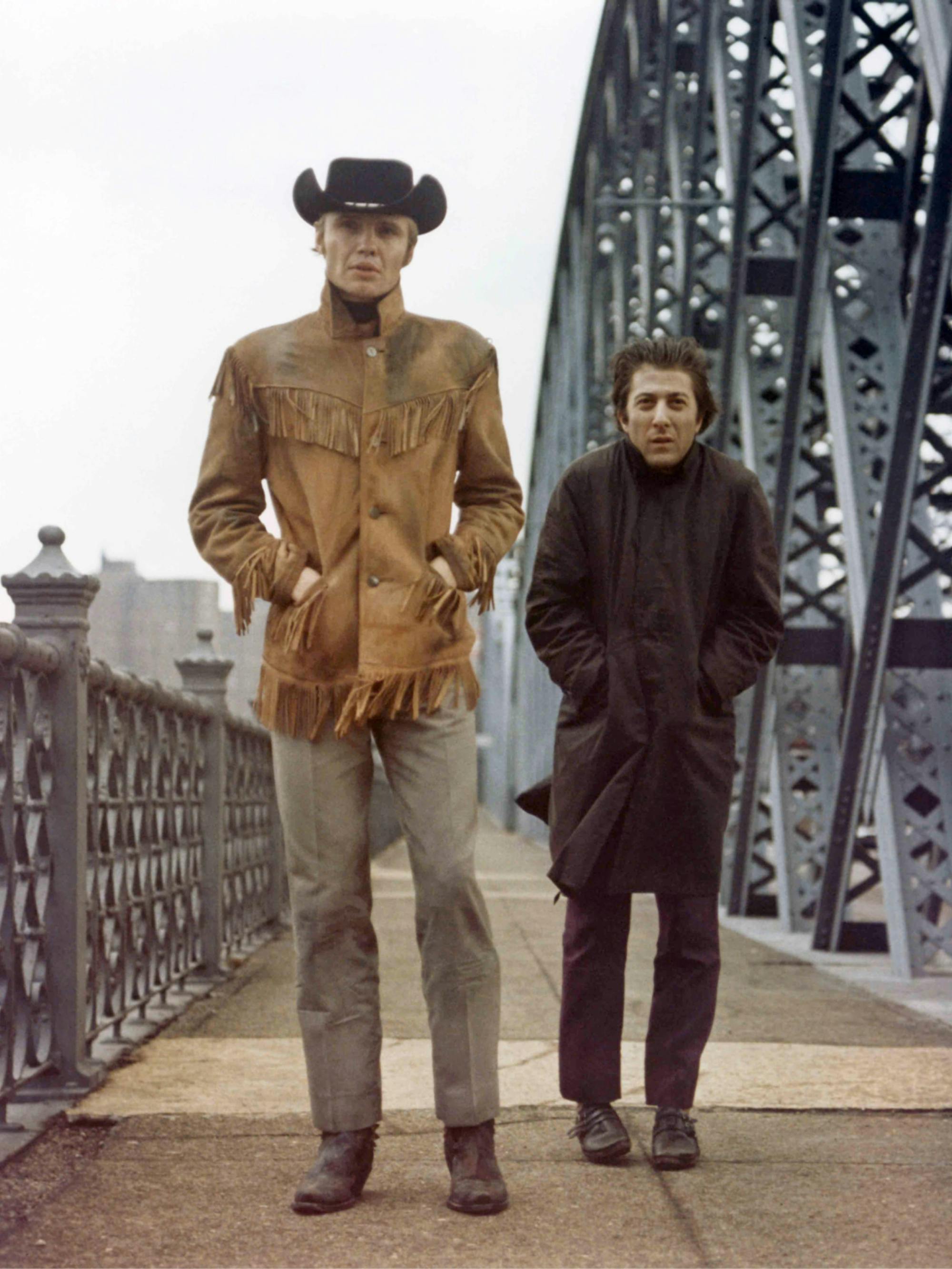 John Voight and Dustin Hoffman walk across a bridge in Midnight Cowboy. Voight wears a fringed jacket and cowboy hat while Hoffman walks a few feet behind him, bracing himself against the cold wind. 