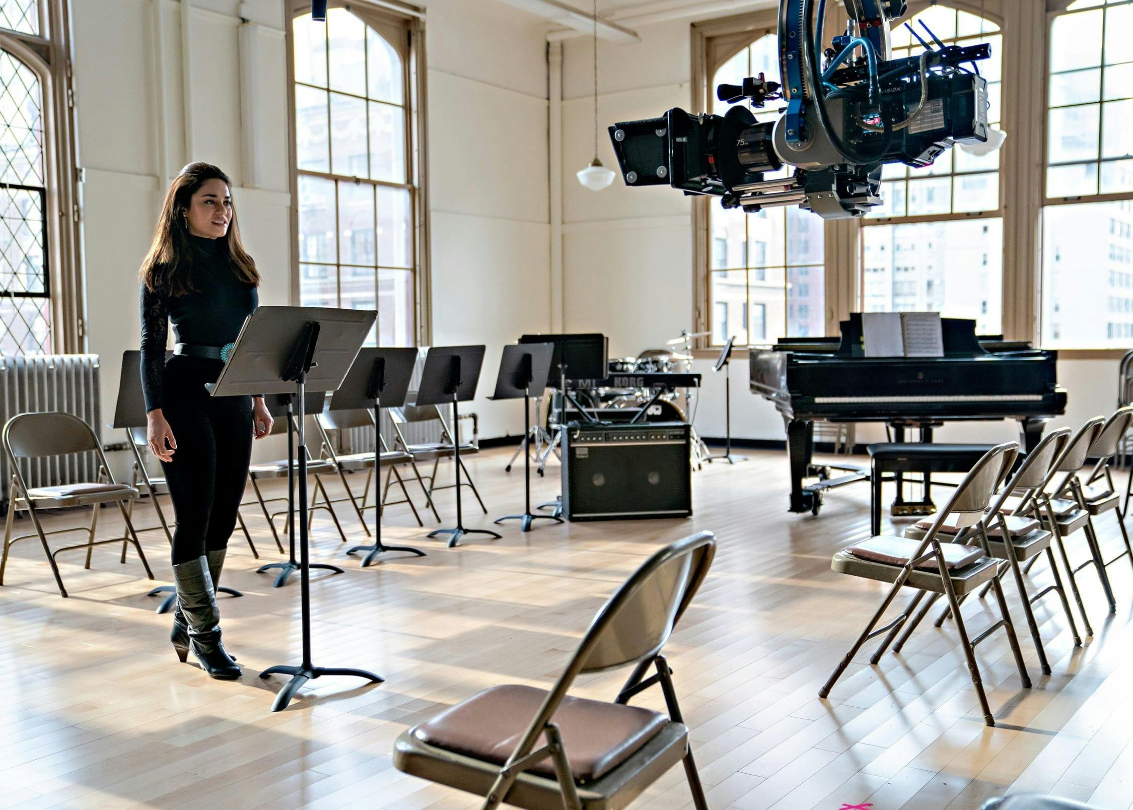 Vanessa Hudgens wears an all-black ensemble and stands behind a music stand in an empty room.