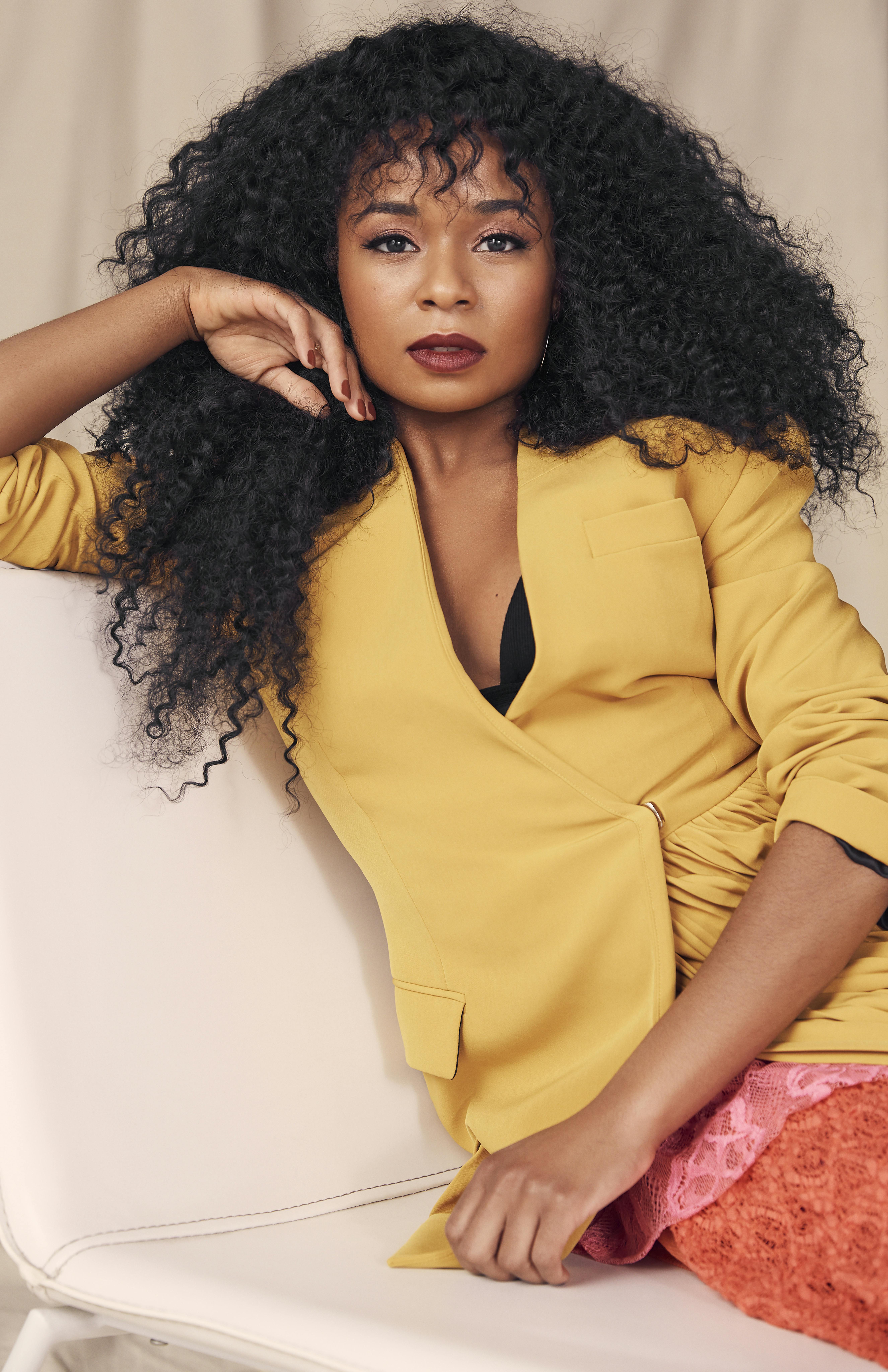 Alexis Floyd sits on an off-white chaise lounge wearing a yellow blazer and lace skirt with pink and orange panels. Floyd's right hand is gracefully framing her face and left hand is seen draped over her lap.