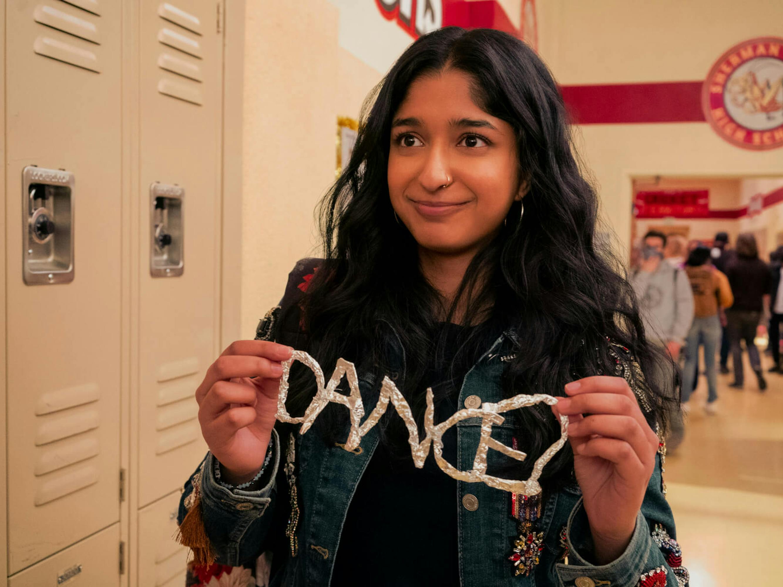 Devi Vishwakumar (Maitreyi Ramakrishnan) wears a jean jacket and holds up the word ‘dance’ made out of tinfoil and smiles sheepishly by the lockers. A classic high school scene!