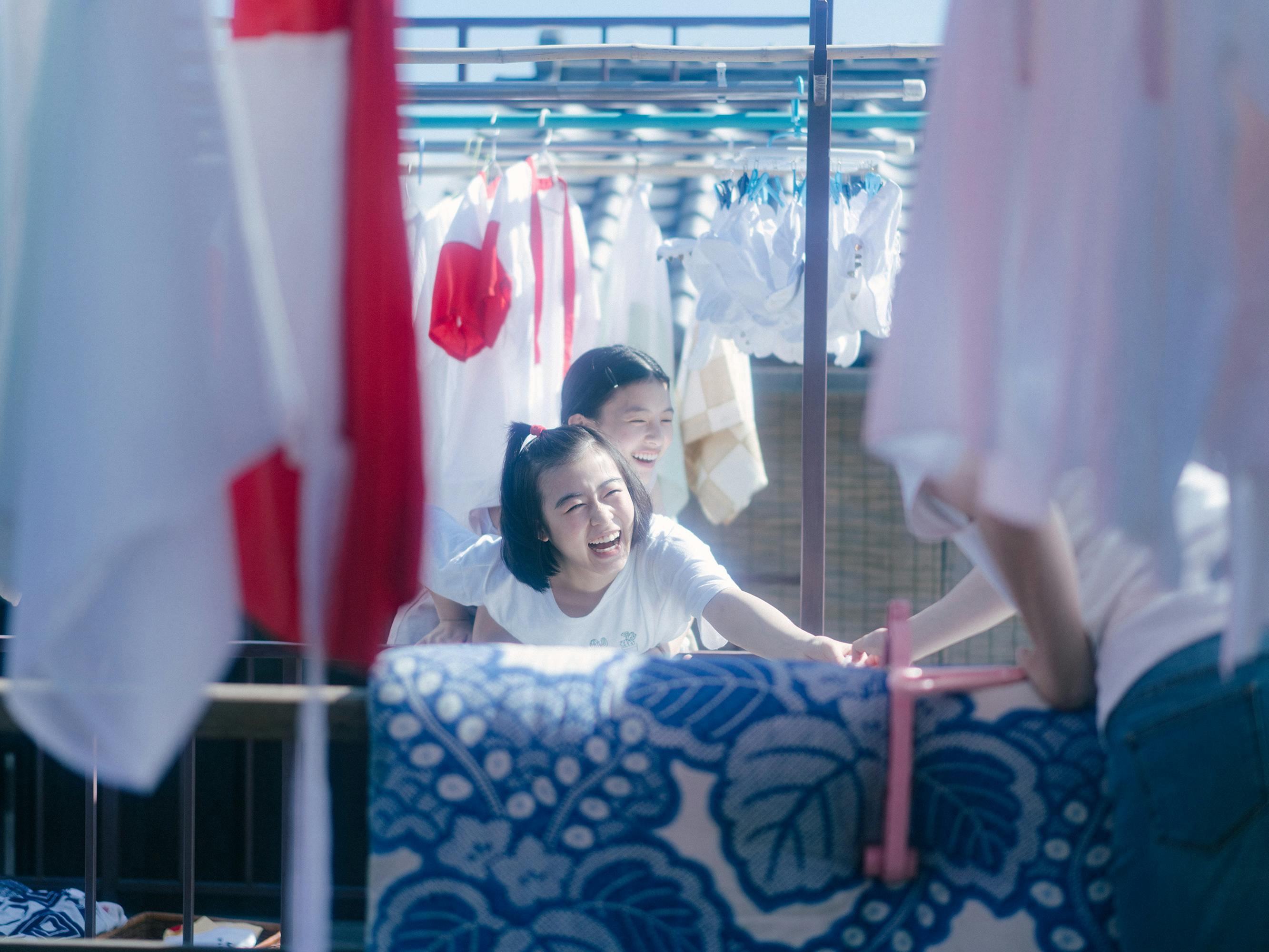 Kiyo (Nana Mori) and Sumire (Natsuki Deguchi) laugh together in the bright light. Behind them are some clothes hanging on hangers. 