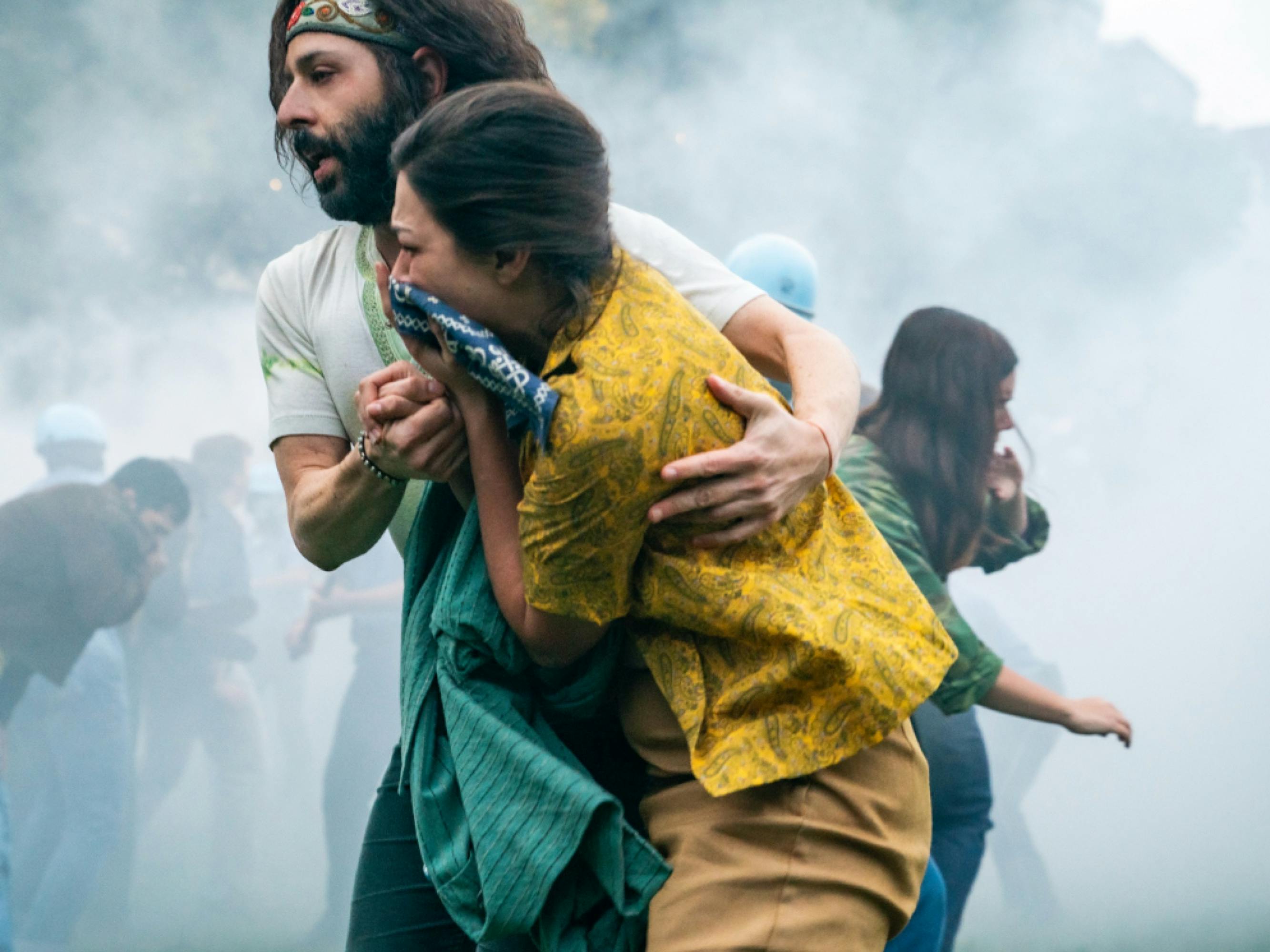 In a still from The Trial of the Chicago 7, Jerry Rubin (Jeremy Strong) and a protestor make their way through a cloud of tear gas. Rubin has his arm around the protestor, who is holding a handkerchief to her mouth and nose. 
