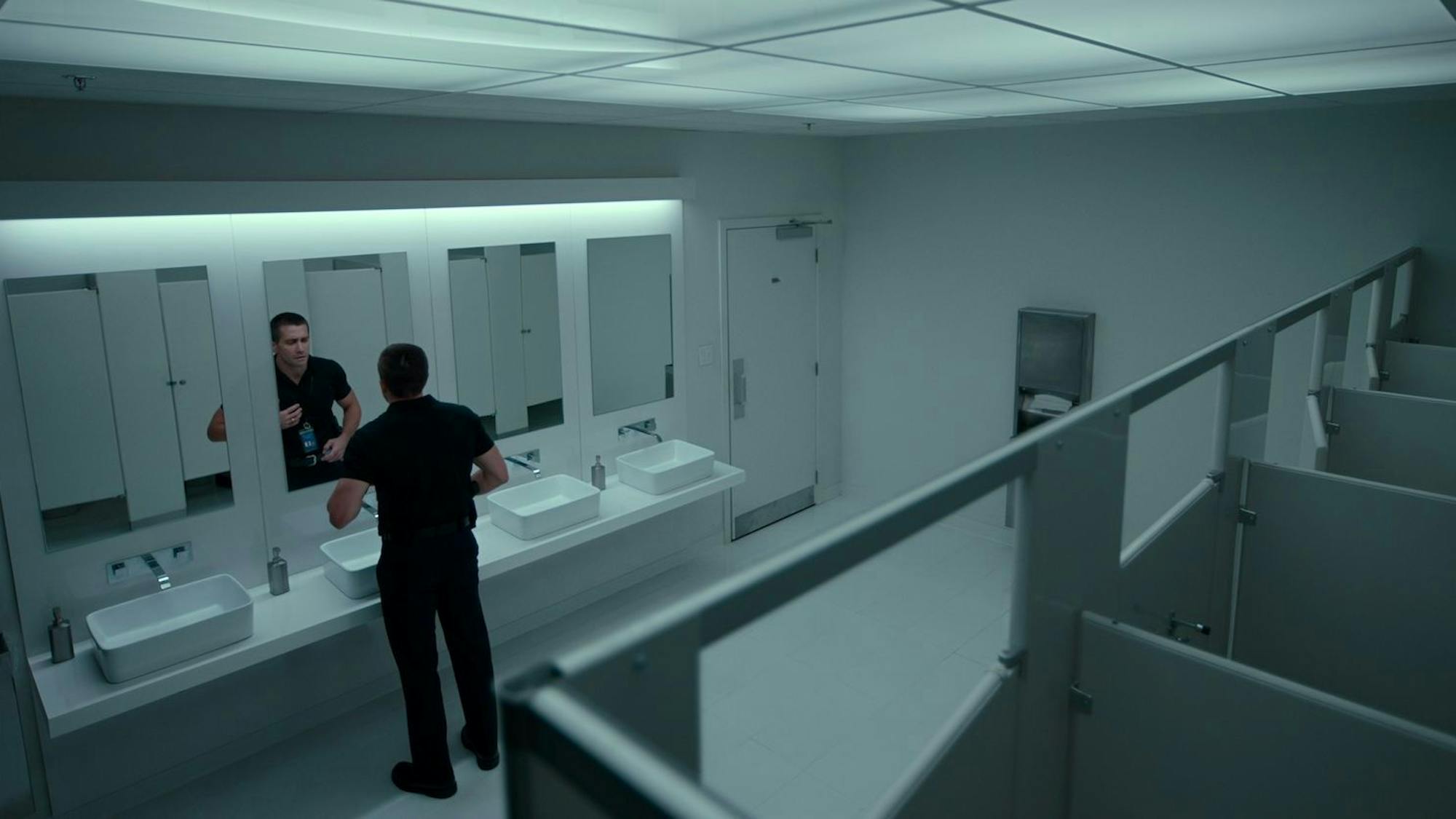 Joe Baylor wears a dark outfit that contrasts with the all white bathroom in which he examines himself in the mirror. 