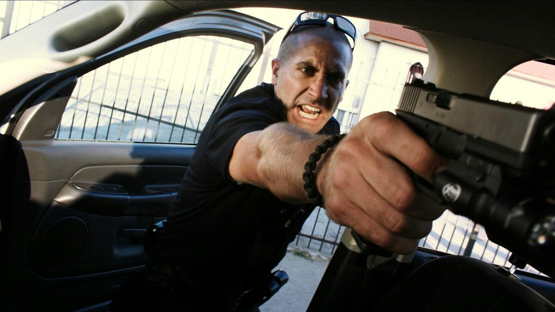 Brian Taylor (Jake Gyllenhaal) in End of Watch (2012) wears all black, sunglasses, and a dark bracelet in this intense, close-up shot. He brandishes a gun towards someone offscreen.