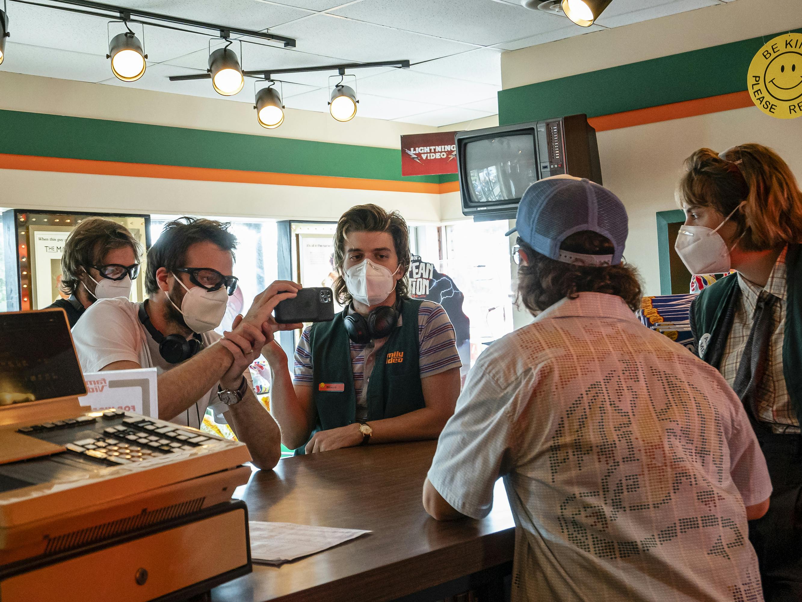 Matt Duffer, Ross Duffer, Joe Keery, Gaten Matarazzo, and Maya Hawke stand around a store counter looking at a phone.