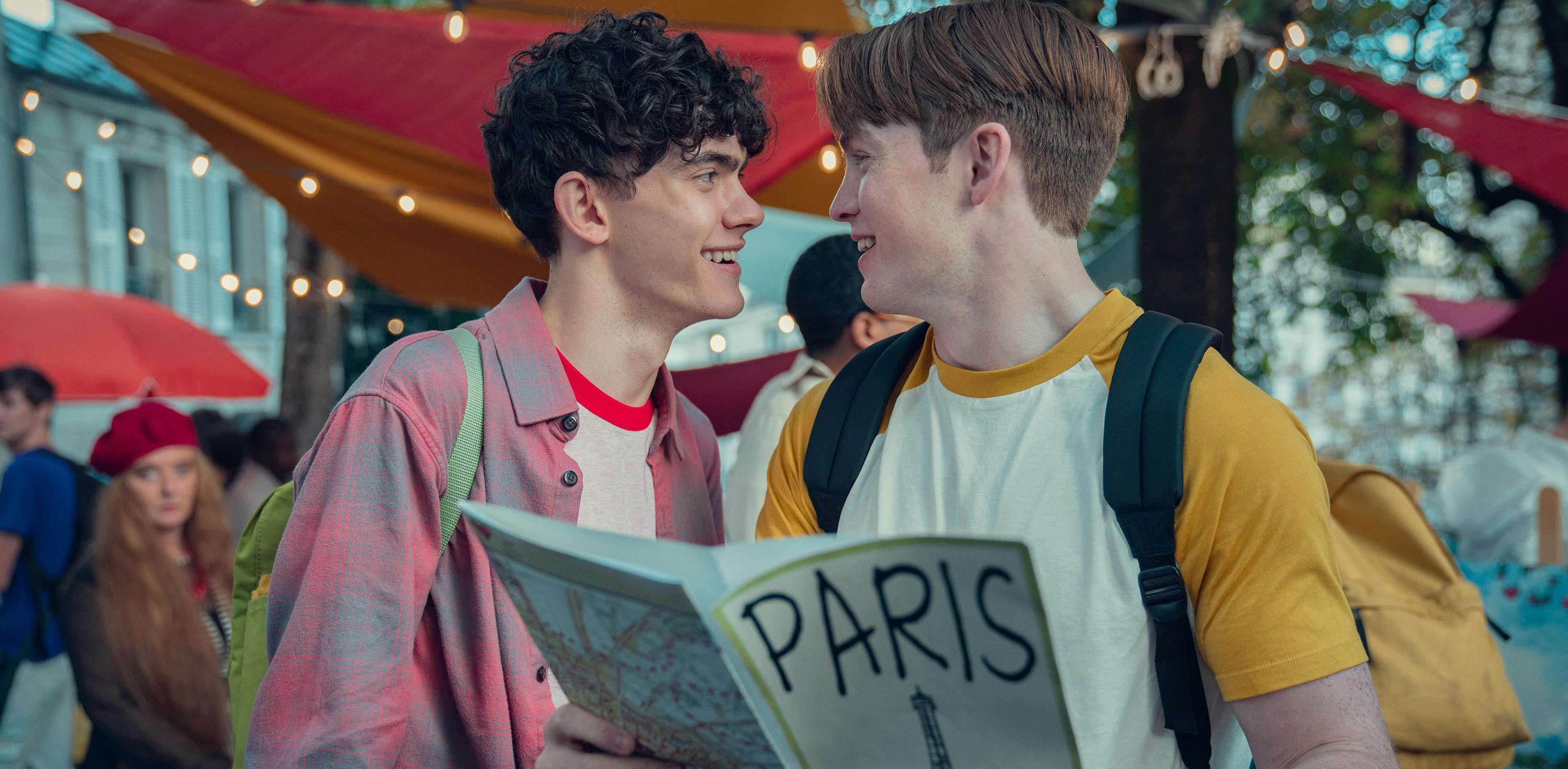 Charlie Spring (Joe Locke) and Nick Nelson (Kit Connor) share a romantic moment over a book titled Paris. Charlie wears a plaid button-up and Nick wears a yellow-and-white baseball shirt.