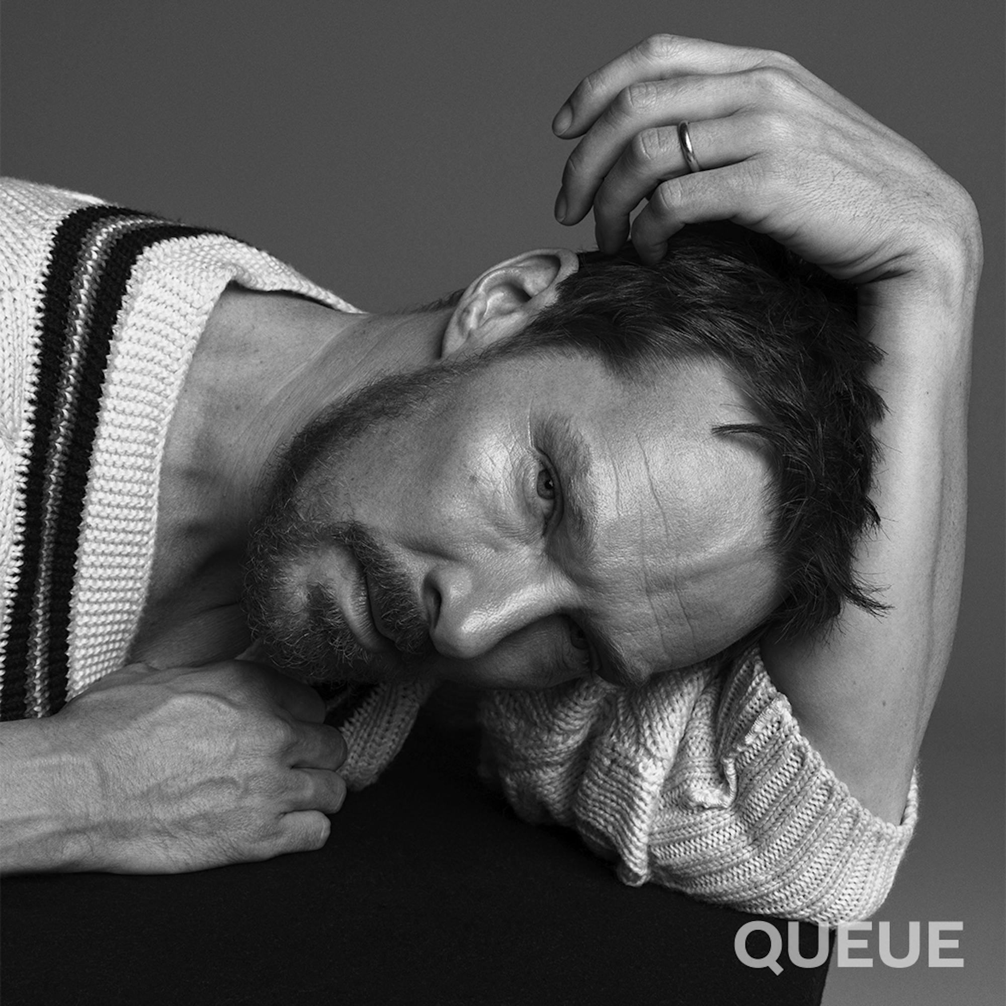 Benedict Cumberbatch wears a white cable knit sweater and rests his head in the crook of his arm. 