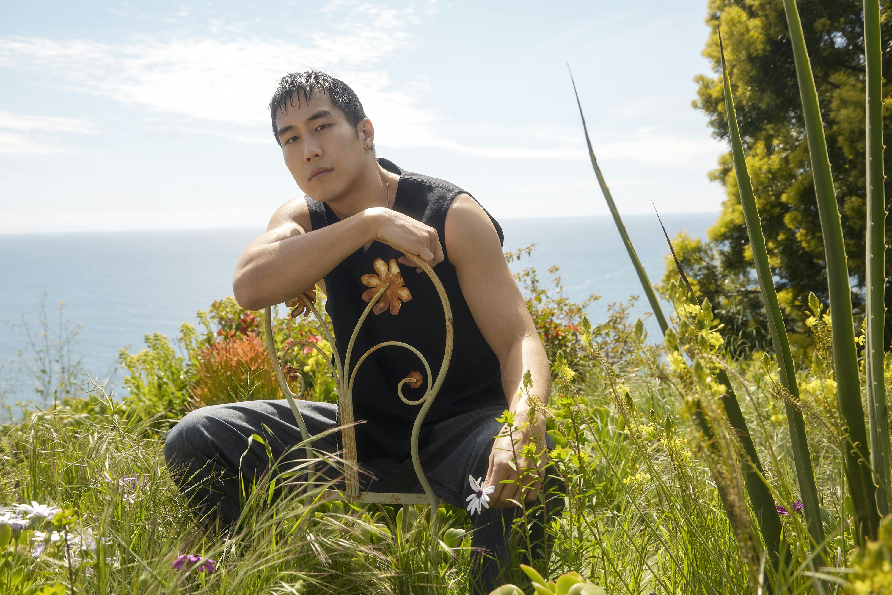 Young Mazino looks like the original man in this sunny, verdant shot. He sits on a curlicue metal chair and wears all black—including a black tank top. His hair is combed forward and he smizes at the camera. 