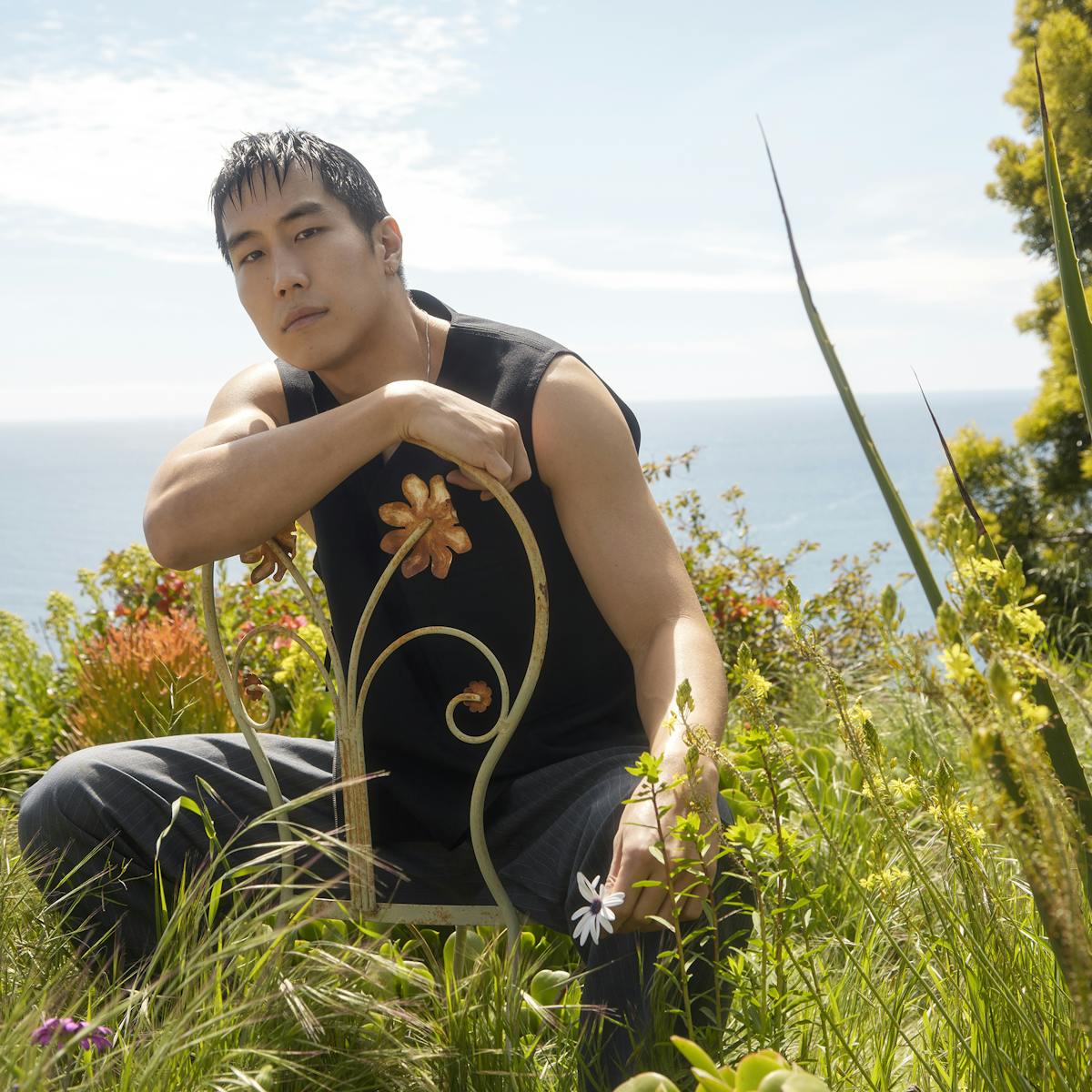 Young Mazino looks like the original man in this sunny, verdant shot. He sits on a curlicue metal chair and wears all black—including a black tank top. His hair is combed forward and he smizes at the camera. 