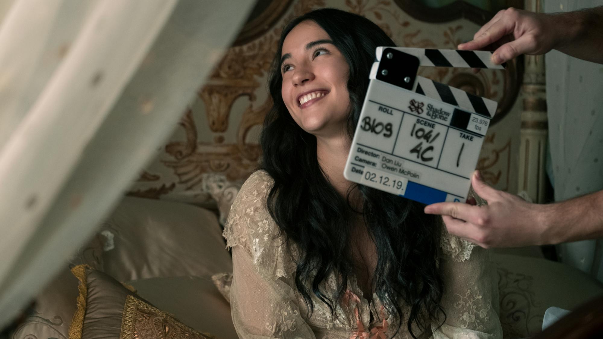 Mei Li sitting on a bed with a clapperboard in front of her face. Her hair is loose, and she wears a white lacy dress. This shot is a break from the tough warrior looks she’s normally seen spotting.