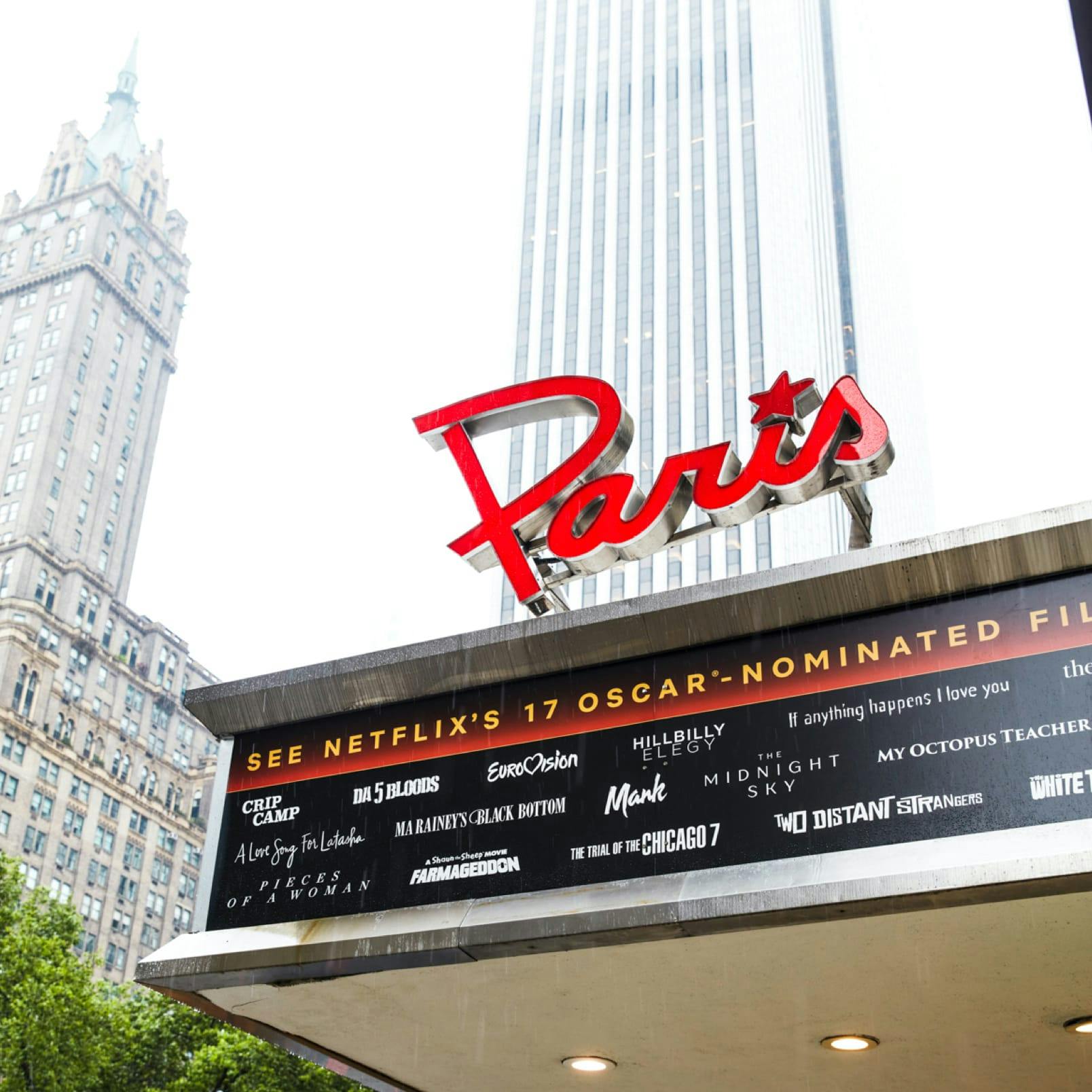 The outside of the Paris Theater. In the background is the plaza hotel, a silvery high rise building, and some trees. Names of Netflix films are written in white on a black marquee, and the Paris head is in red, vintage font.