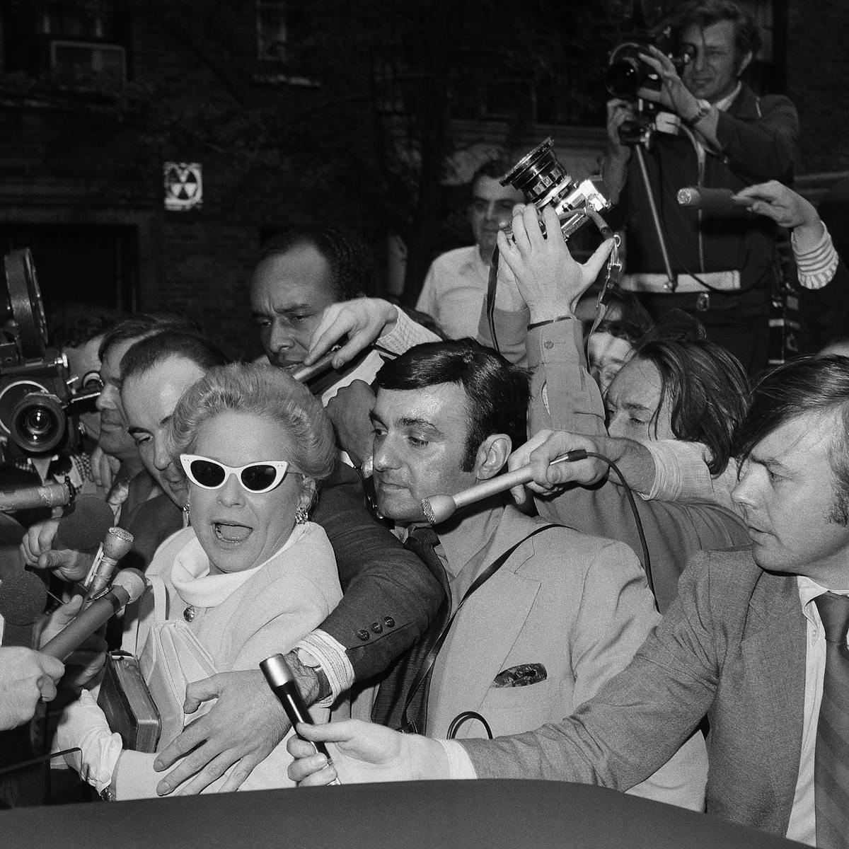 Martha Mitchell wears some white cat-eye glasses and fights her way through a crowd of press.