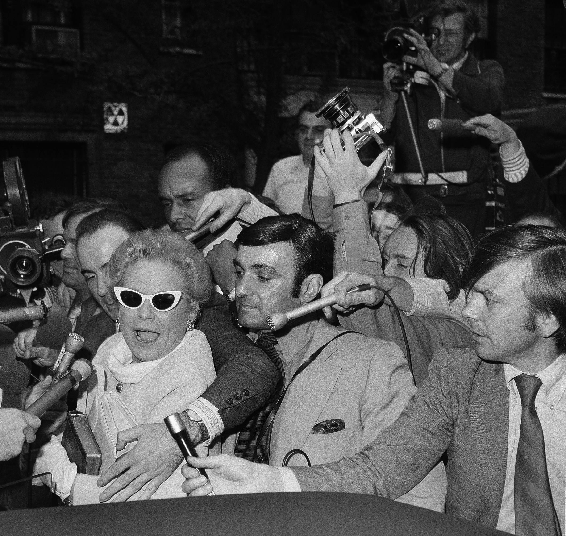 Martha Mitchell wears some white cat-eye glasses and fights her way through a crowd of press.