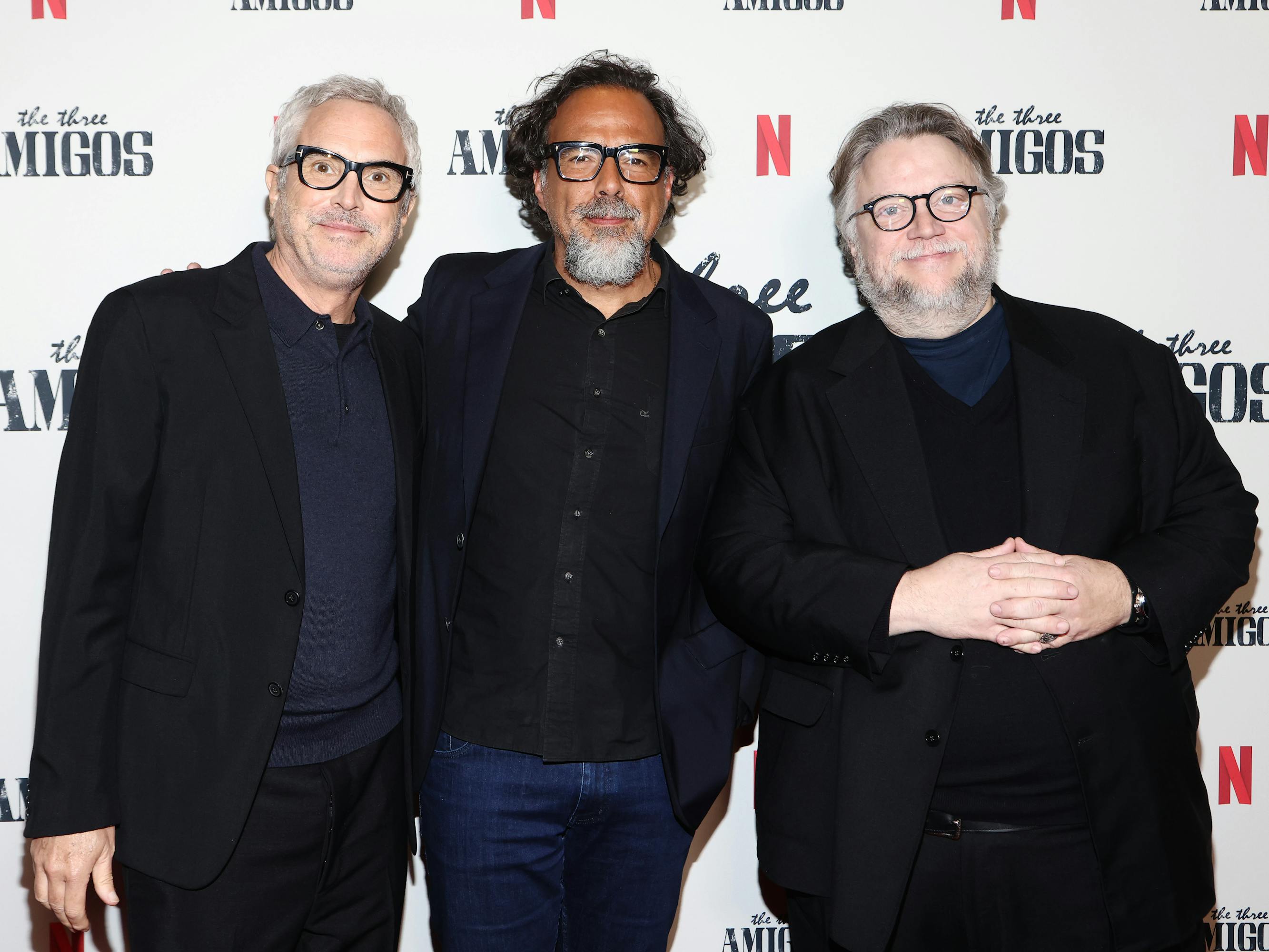 Alfonso Cuarón, Alejandro Iñárritu, and Guillermo del Toro stand together on a red carpet wearing black glasses in various degrees of chunkiness. The step-and-repeat background features the Netflix icon, and 'the three Amigos' in faded black font.