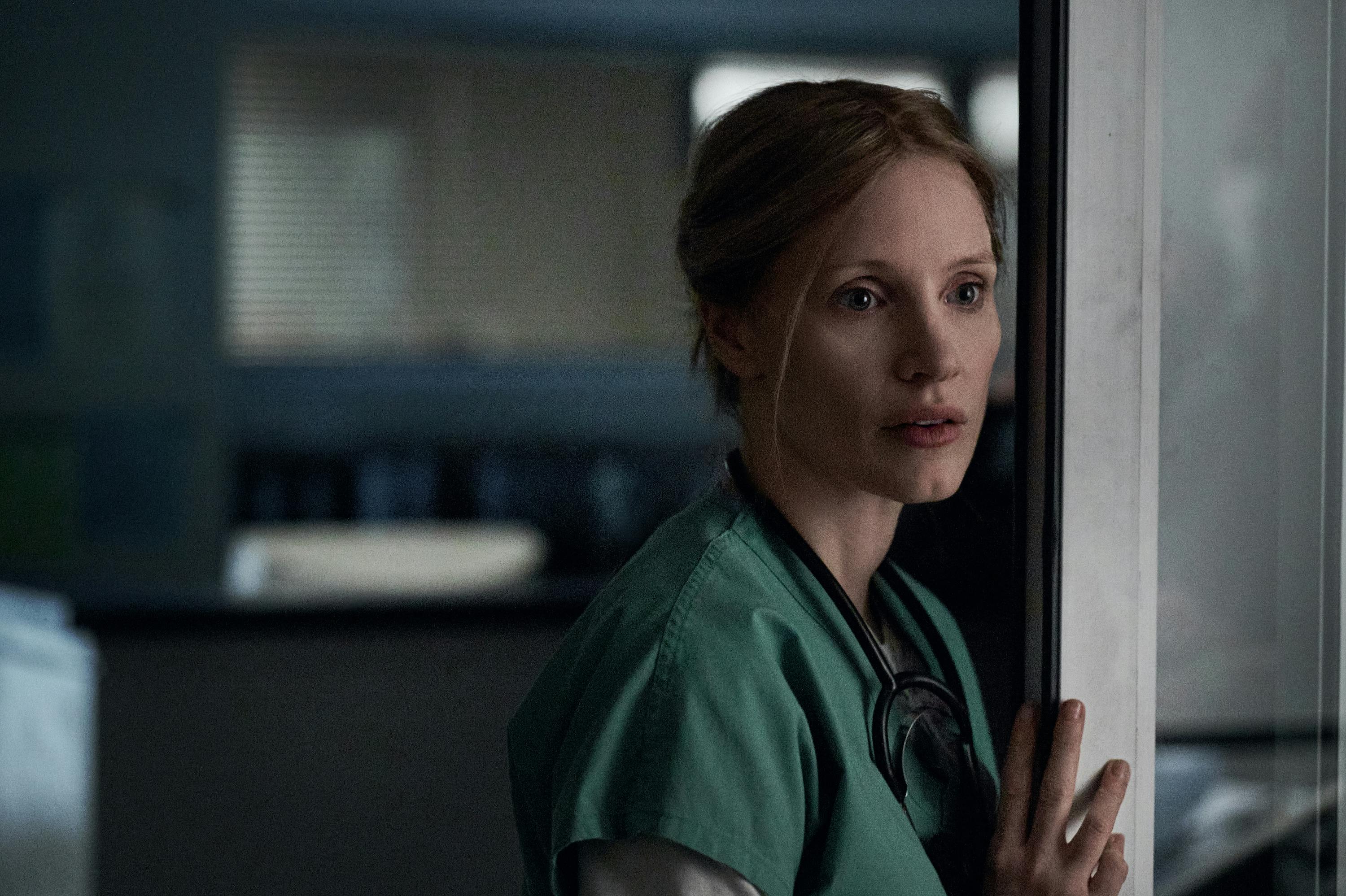 Jessica Chastain wears green scrubs and leans on a grey wall.