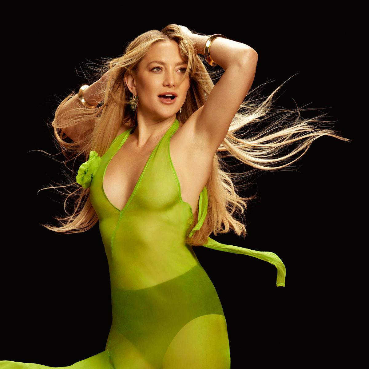 Kate Hudson wears a green dress and black underwear, striking a pose against a black background.
