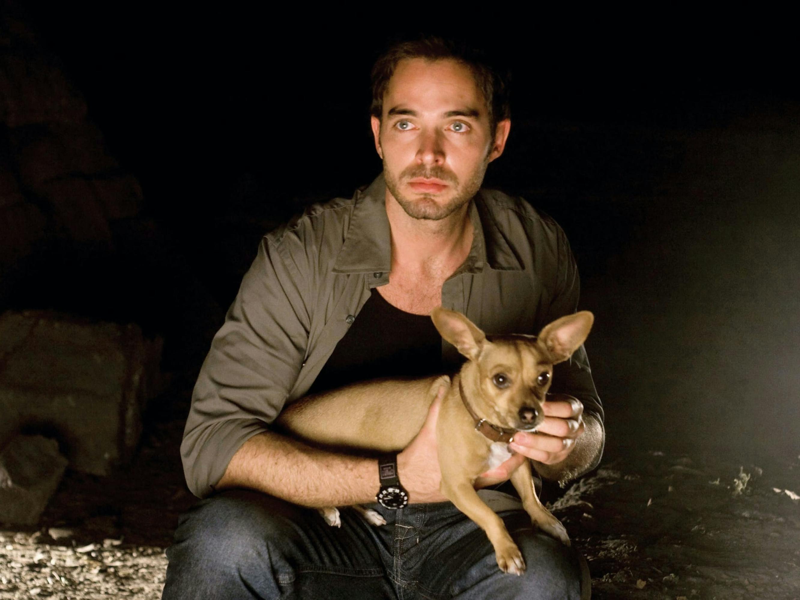 Álex (Manolo Cardona) holds a light brown chihuahua in his arms. The scene is dark, but his figure is illuminated. He wears a green long sleeved shirt and jeans and looks intensely at something off screen.