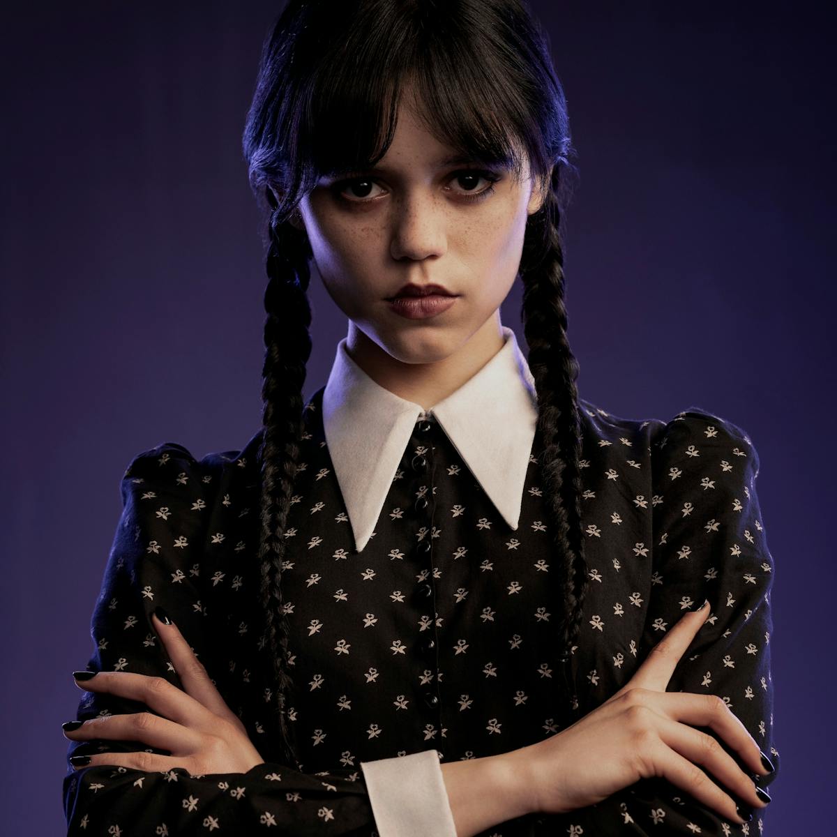 Wednesday Addams (Jenna Ortega) wears a black dress with white polka dots and a triangular white collar and two braids against a purple background.