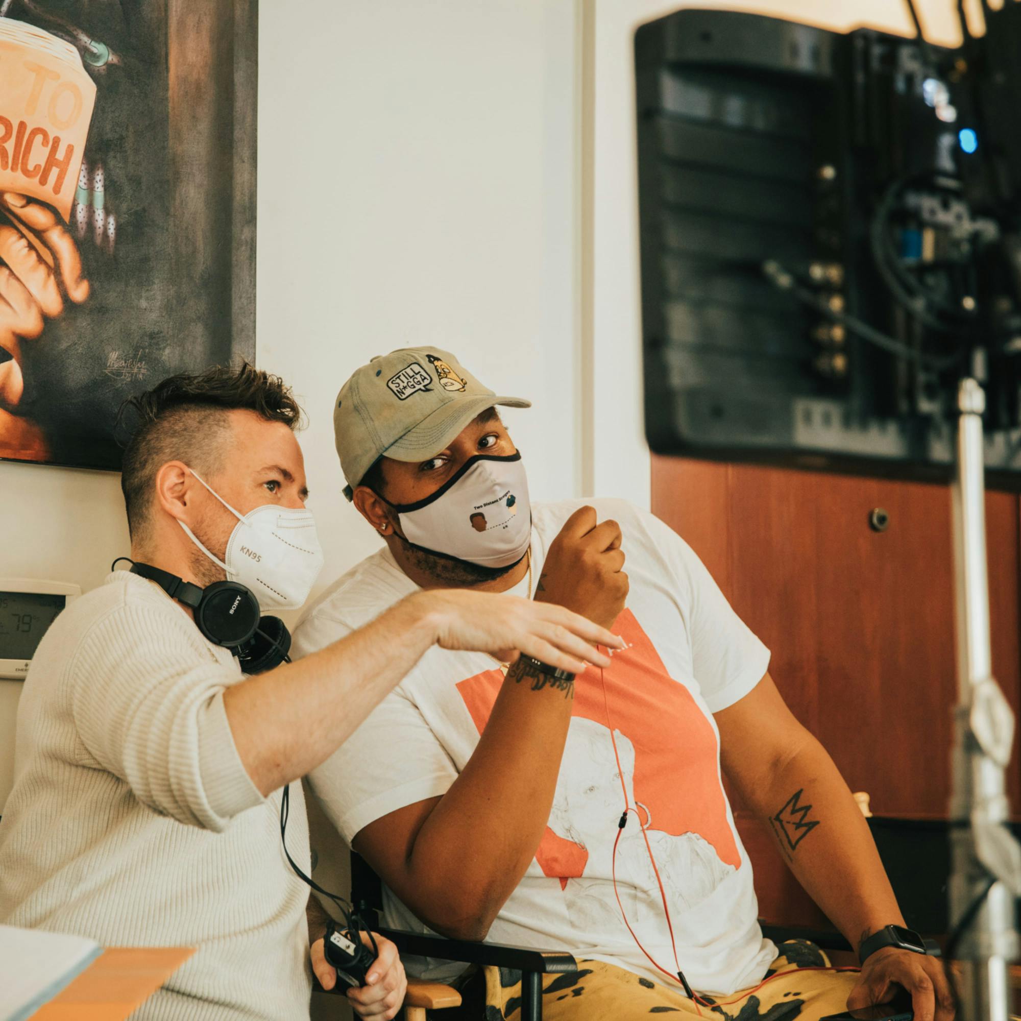 Directors Martin Desmond Roe and Travon Free from left to right. Roe wears a gray sweater with headphones around his neck, and a white mask. Free also wears a white mask, with a tan hat and a white and orange shirt. The shot is candid and the two men discuss something, although Free seems to be aware that someone is taking a picture of them.