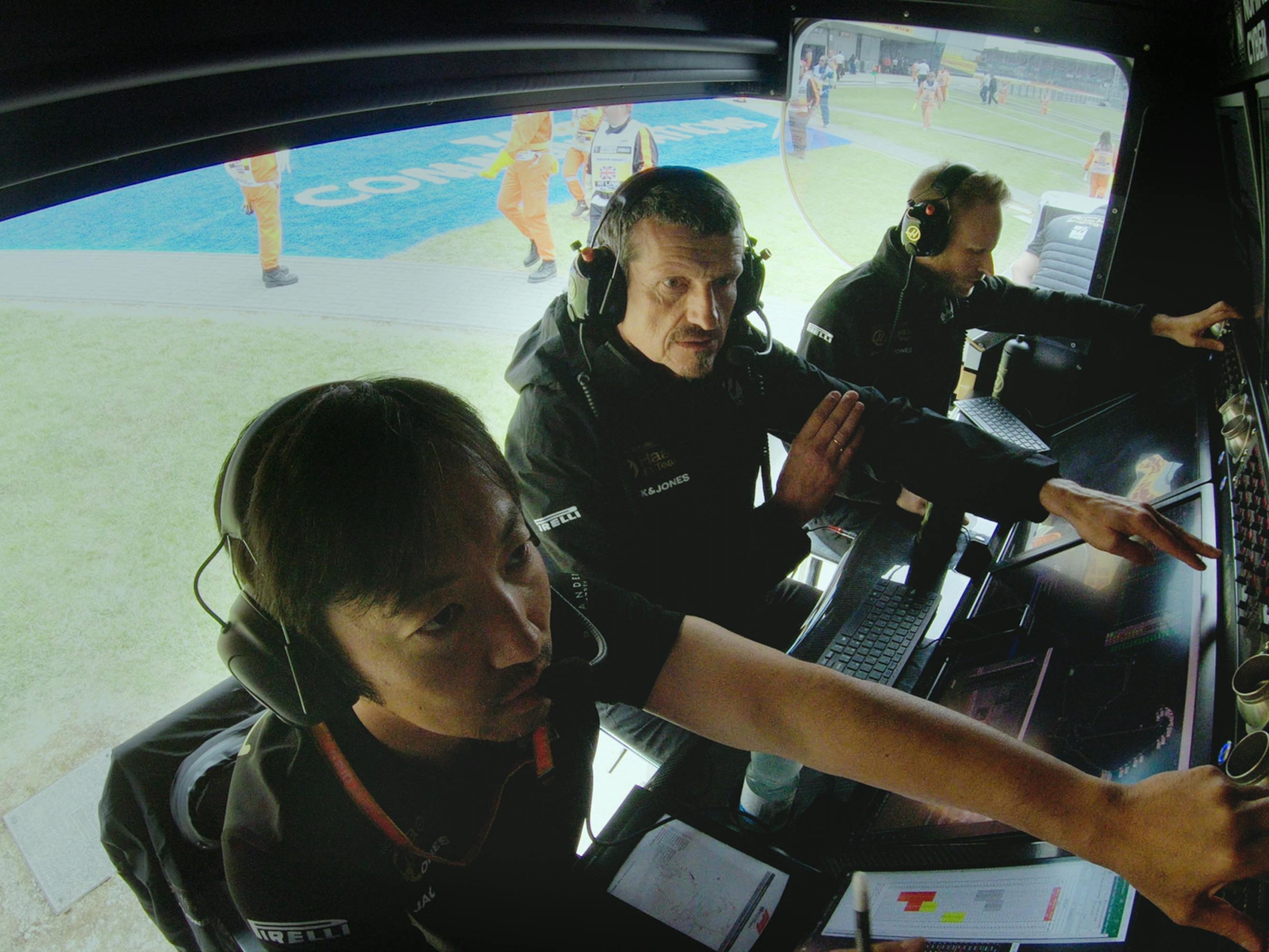 Guenther Steiner sits at a panel of screens, wearing a headset with a mouthpiece, in between two colleagues as they watch the race.