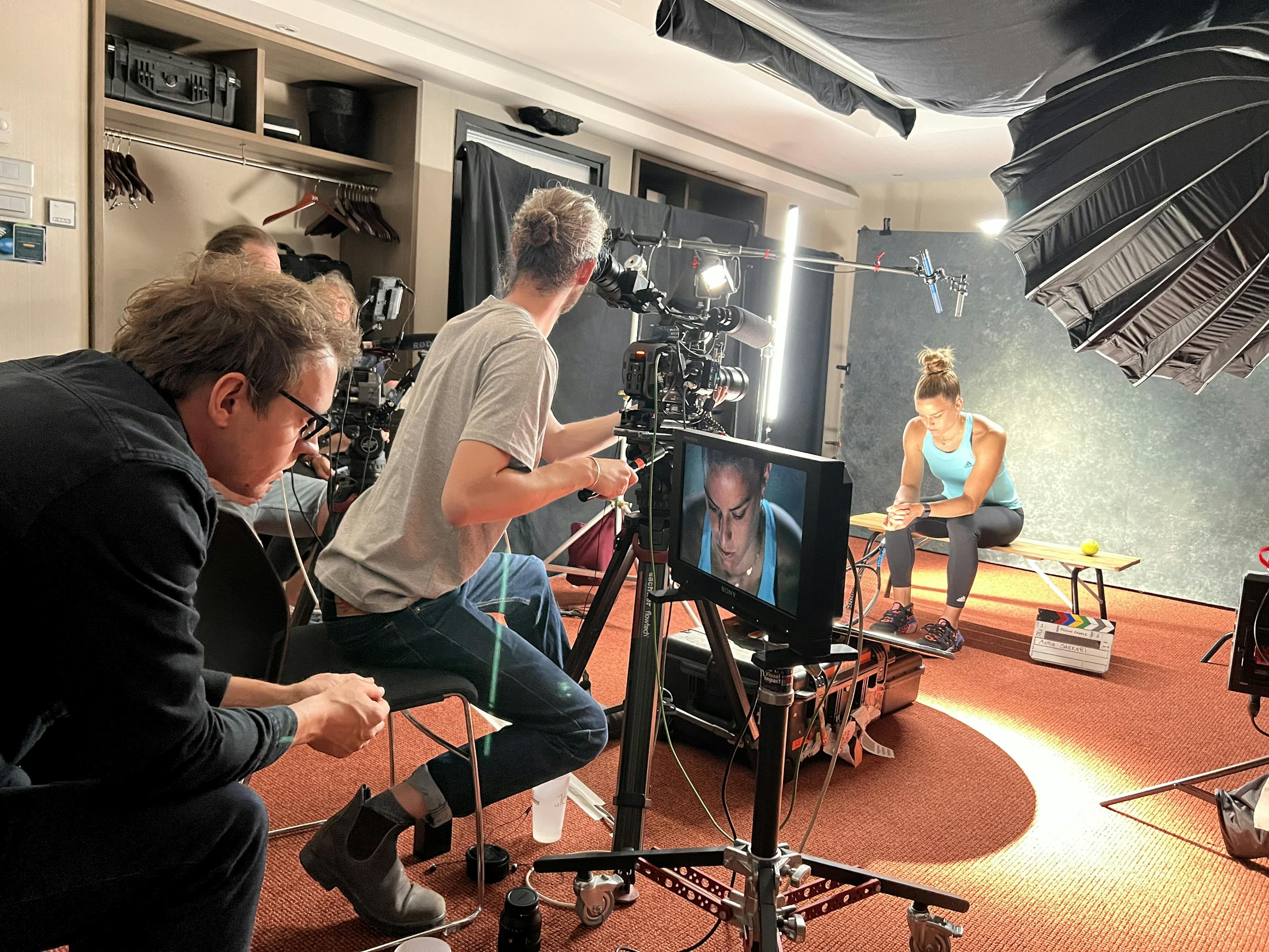 A tennis player sits on a bench in a film studio, waiting to be interviewed. She's surrounded by a production team, cameras, and other film equipment.