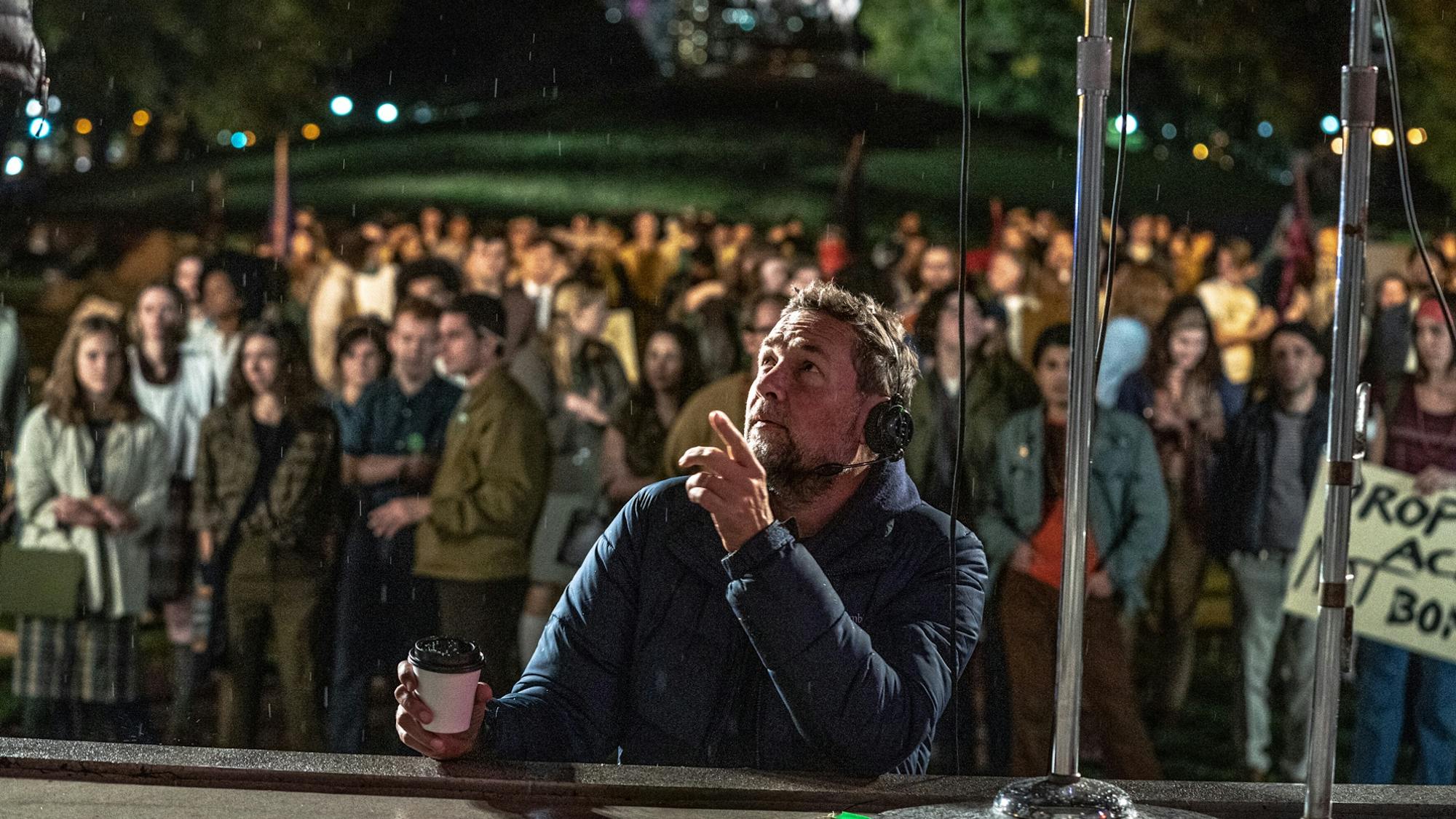 It’s a night shoot in Chicago, and Phedon Papamichael is pictured leaning against a stage where the protest organizers in the film will rally their crowd. He holds a coffee in one hand and points up at something out of the frame with another. Behind him are the extras assembled for the crowd scene.